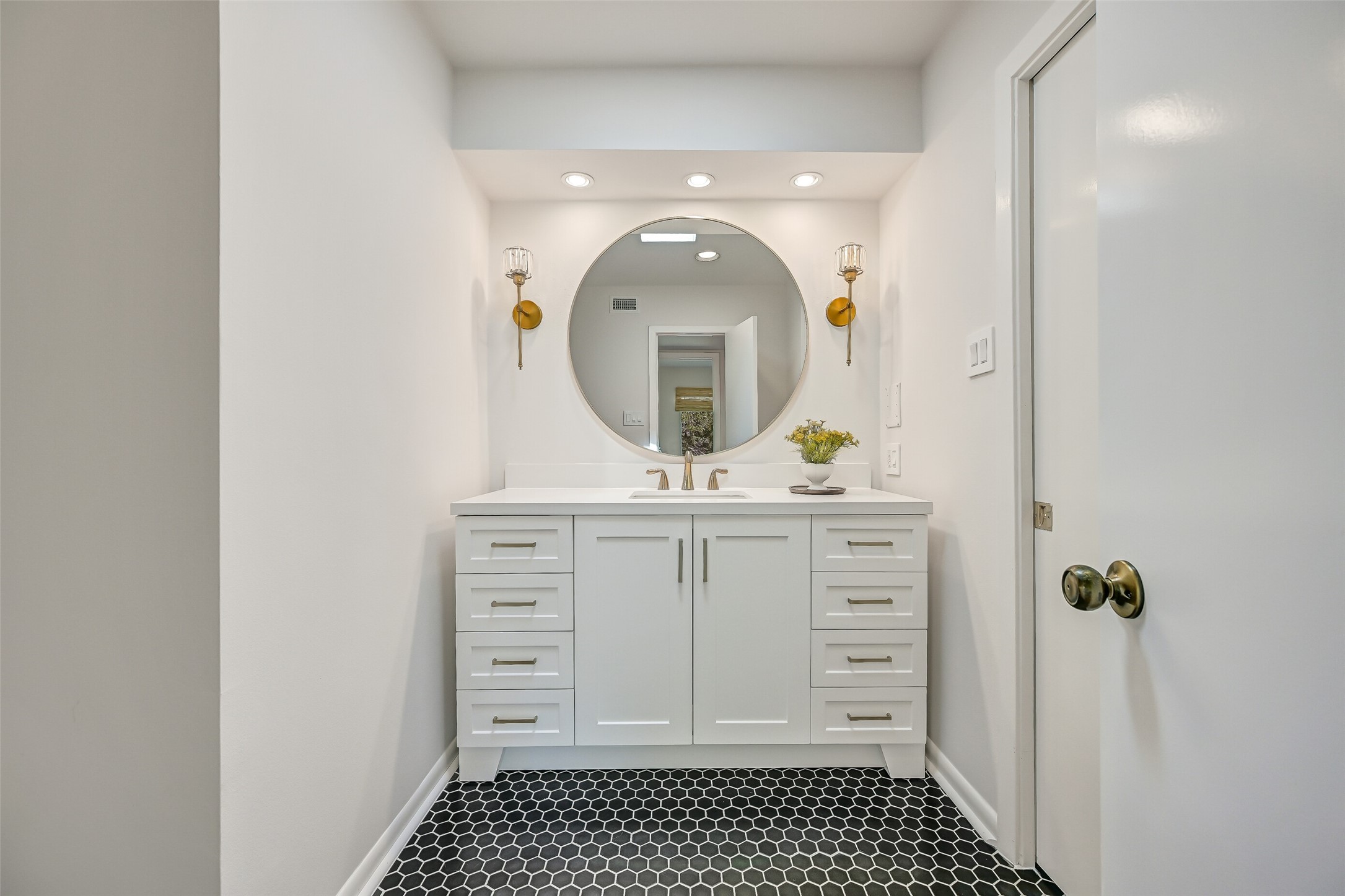 One of two separate vanity areas in the Jack & Jill bathroom, this updated space features a white vanity with generous drawer storage, contemporary hardware, quartz-look countertop & round mirror flanked by decorative gold sconces. Black hexagonal tile flooring adds a striking contrast.