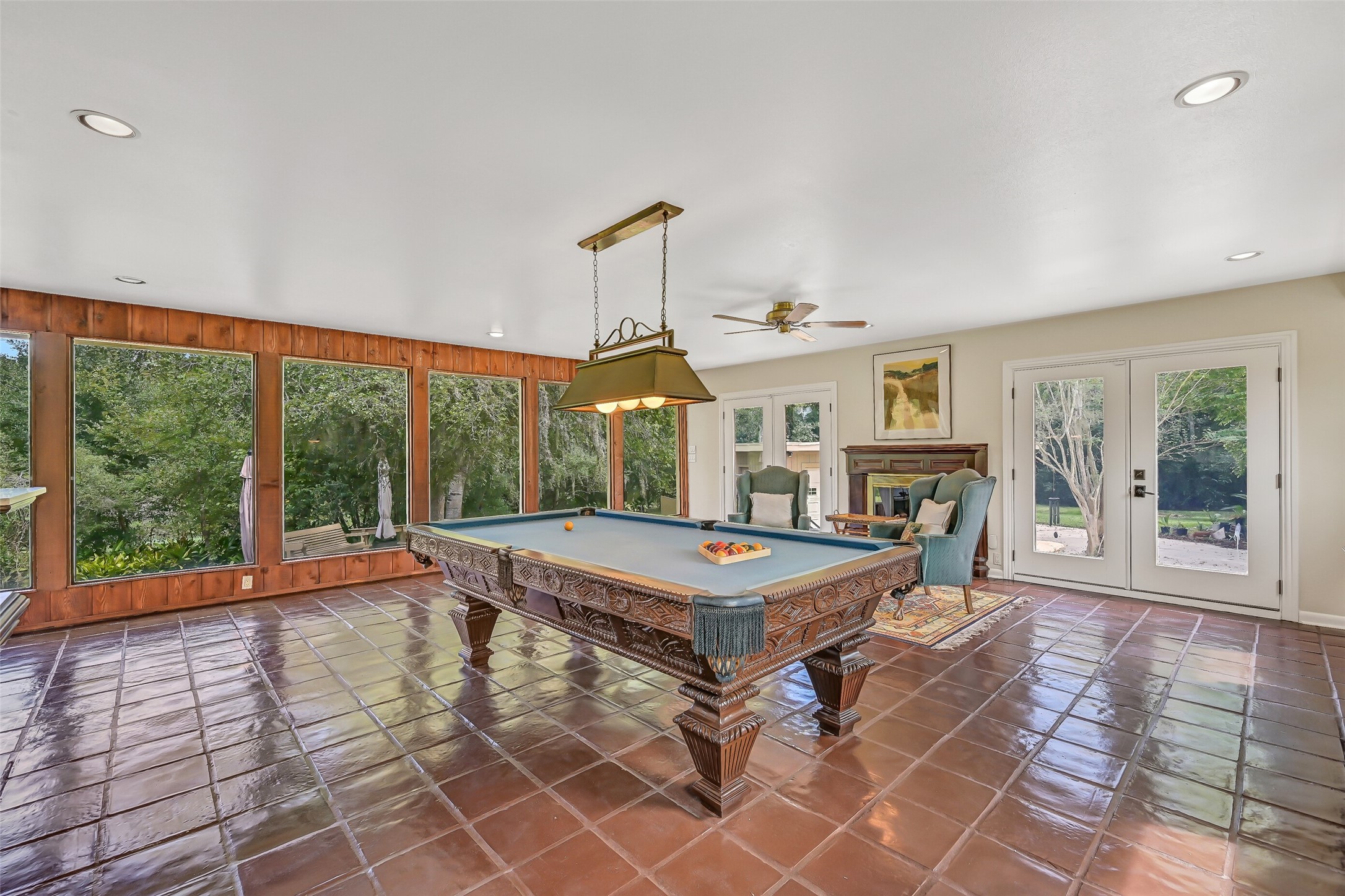 A different perspective showcases the gameroom's beautiful backyard views, with oversized windows framed by rich wood paneling. The pool table sits beneath a decorative hanging light, while a cozy seating area beside the second gas-log fireplace creates another inviting place to gather.
