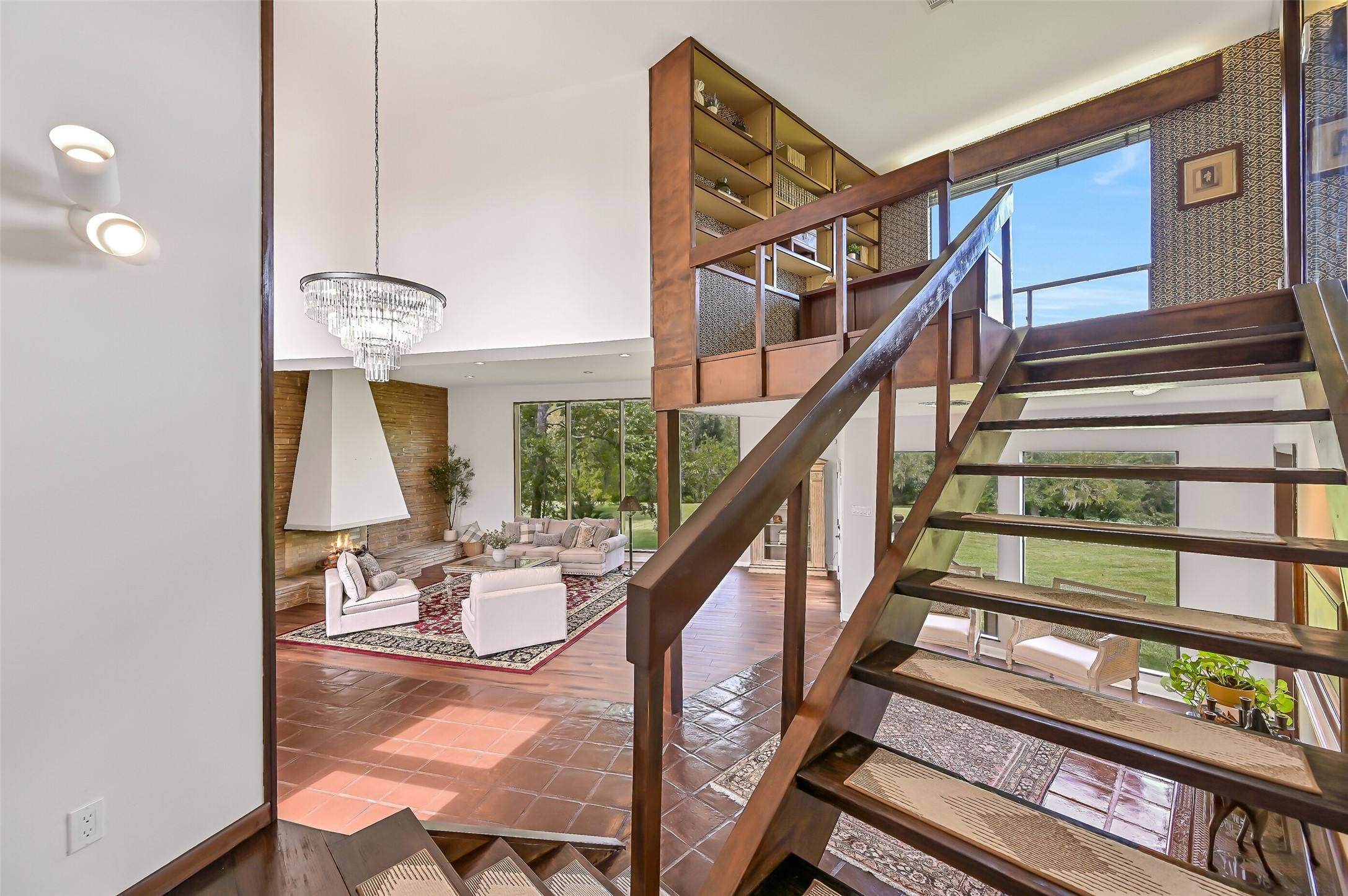 The distinctive open-riser wooden staircase leads from the grand entry to the home's private upper level, reserved for the primary suite & adjoining study. An elevated library with built-in shelving overlooks the living room, while expansive windows & a dramatic chandelier accentuate the open design.