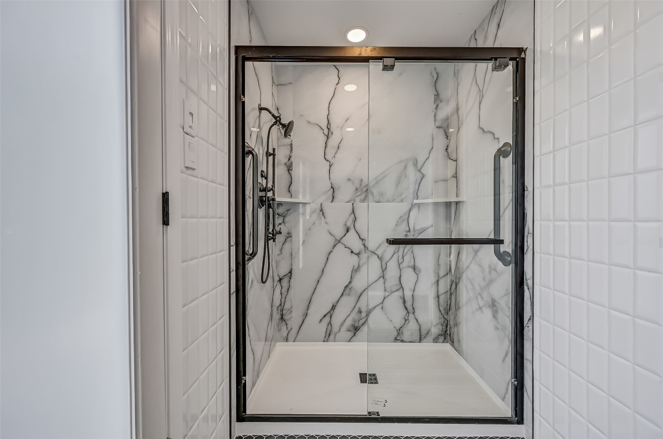 The updated primary shower features dramatic marble-look wall surrounds, a sliding glass enclosure with dark metal framing, built-in corner shelves & a handheld shower fixture. Its generous dimensions provide a comfortable bathing space, complemented by the bathroom's black hexagonal tile flooring.