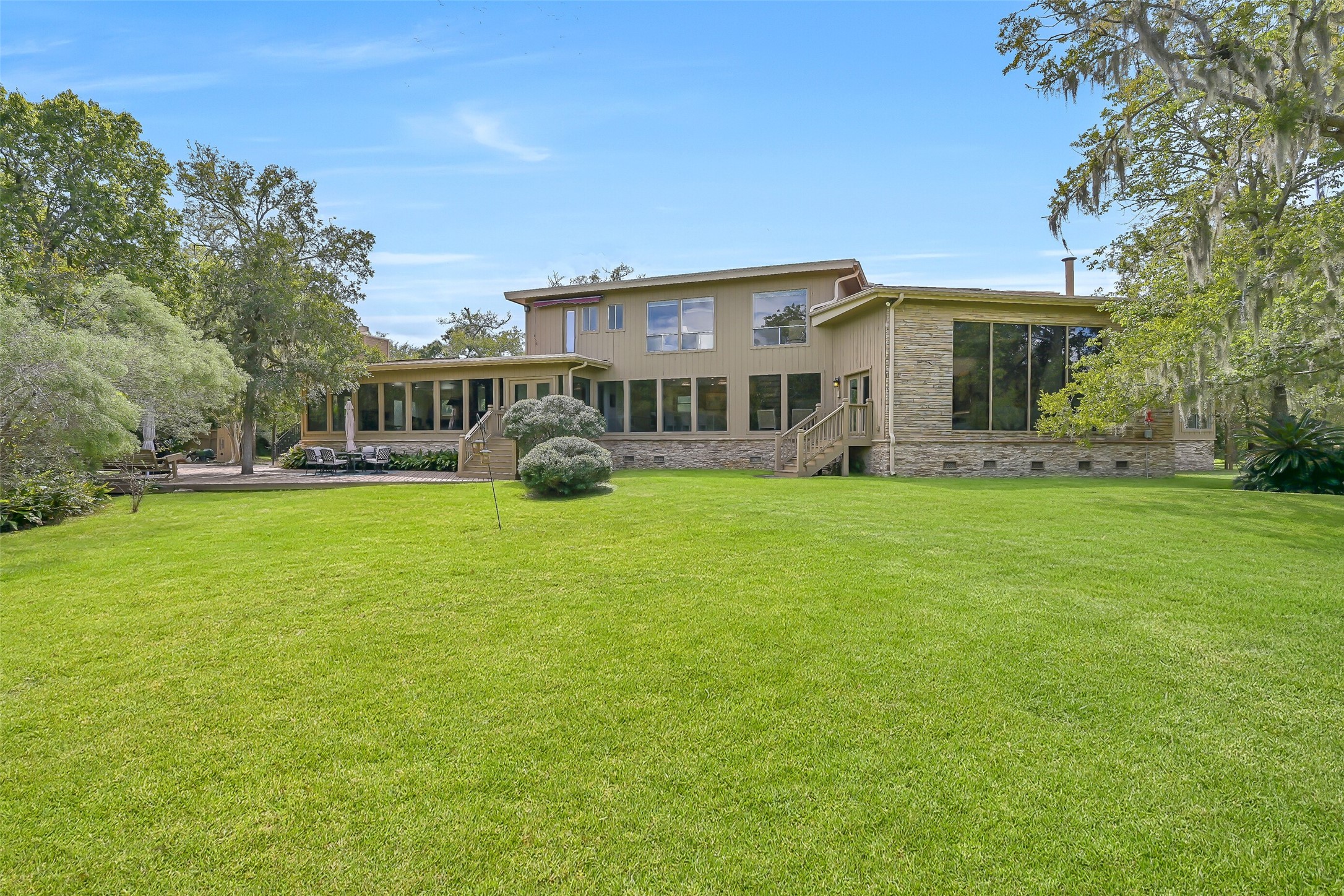 The rear elevation highlights the home's distinctive multi-level architecture, extensive windows & multiple access points to the outdoor living areas. A spacious deck overlooks the expansive grassy backyard, where established trees provide a picturesque backdrop across the acreage.
