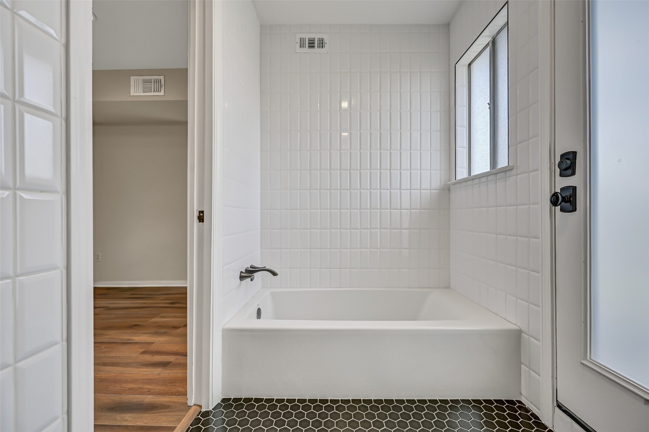 A recently replaced soaking tub sits within a white tiled alcove in the primary bathroom, offering a separate bathing option alongside the updated shower. Black hexagonal tile flooring creates a contrasting finish, while a nearby exterior door provides access to the adjacent roof area.