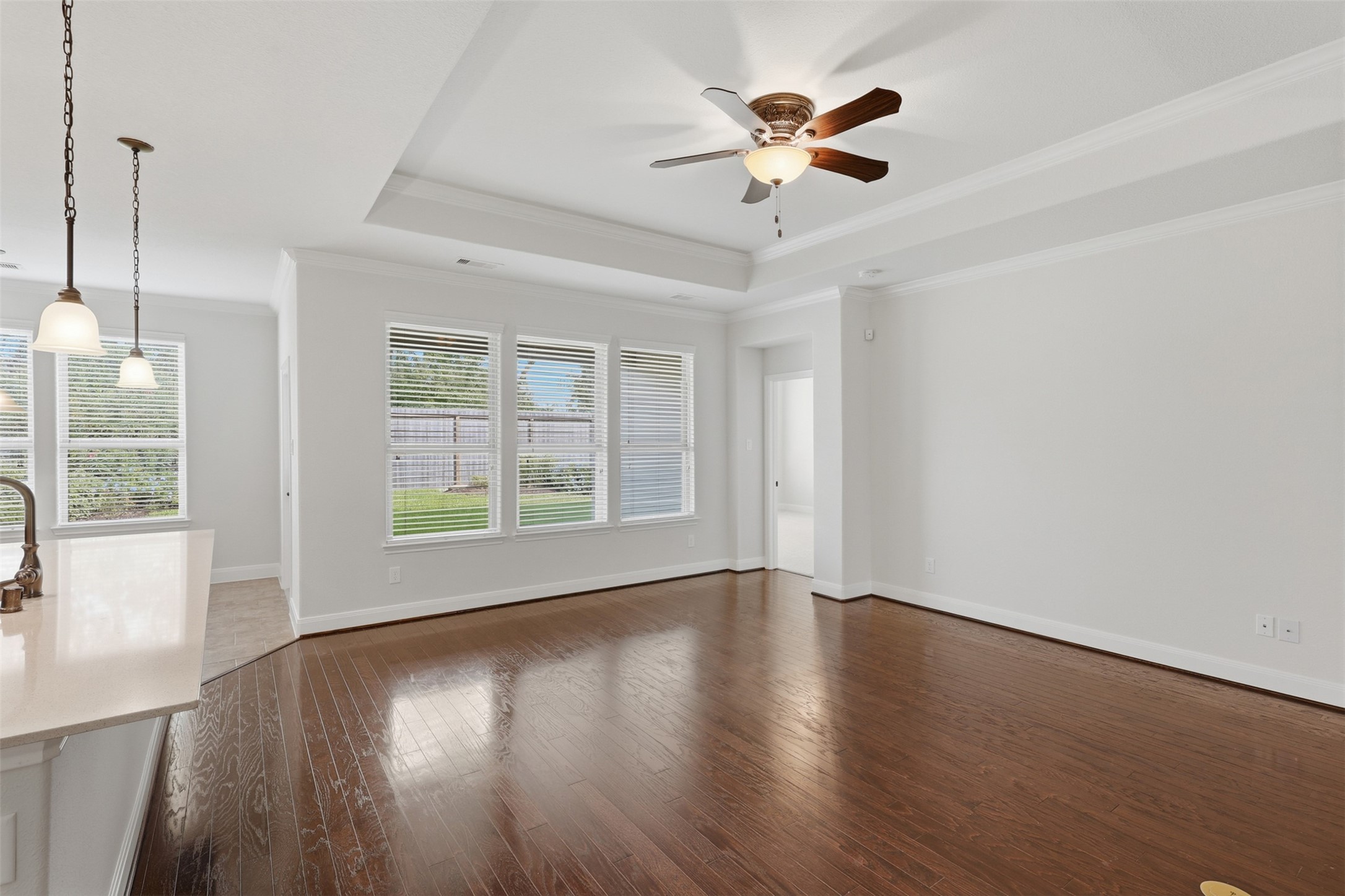 Spacious, light-filled living room featuring beautiful hardwood flooring, crown molding, a tray ceiling, ceiling fan, and a wall of windows overlooking the backyard.