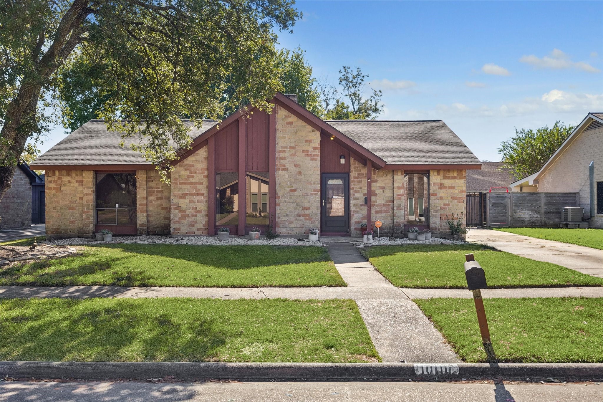 Charming brick home with a unique peaked roof and expansive windows, nestled under the shade of mature trees, offering a welcoming front walkway and lush lawn.