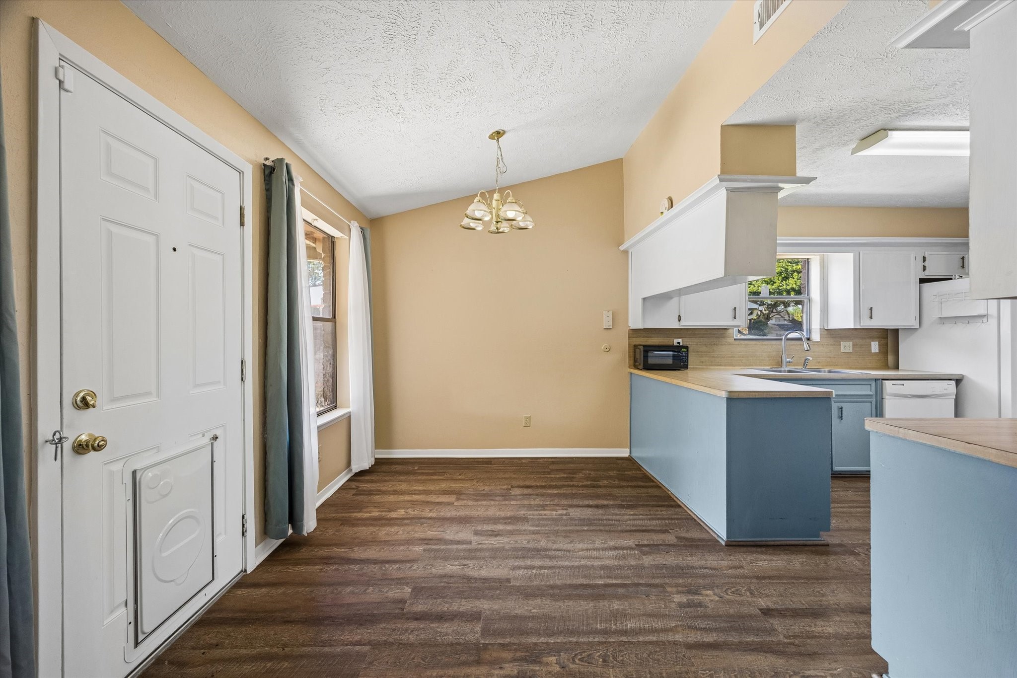 Charming open-concept kitchen and dining area featuring a vaulted ceiling, warm flooring, and ample natural light, with stylish blue cabinetry and a cozy chandelier.