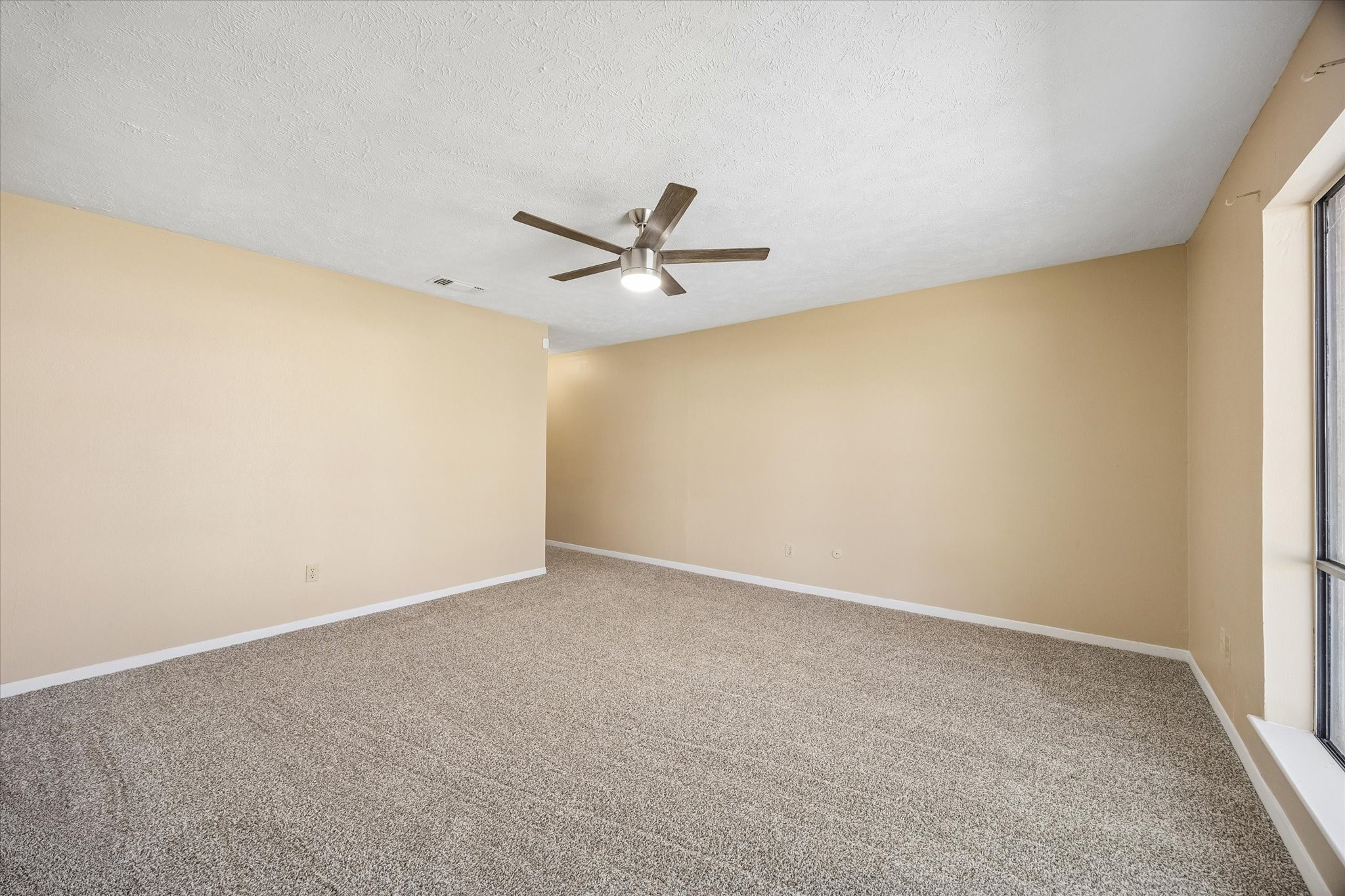 Spacious living area with neutral-toned walls, plush carpeting, and a modern ceiling fan, offering ample natural light through large windows.