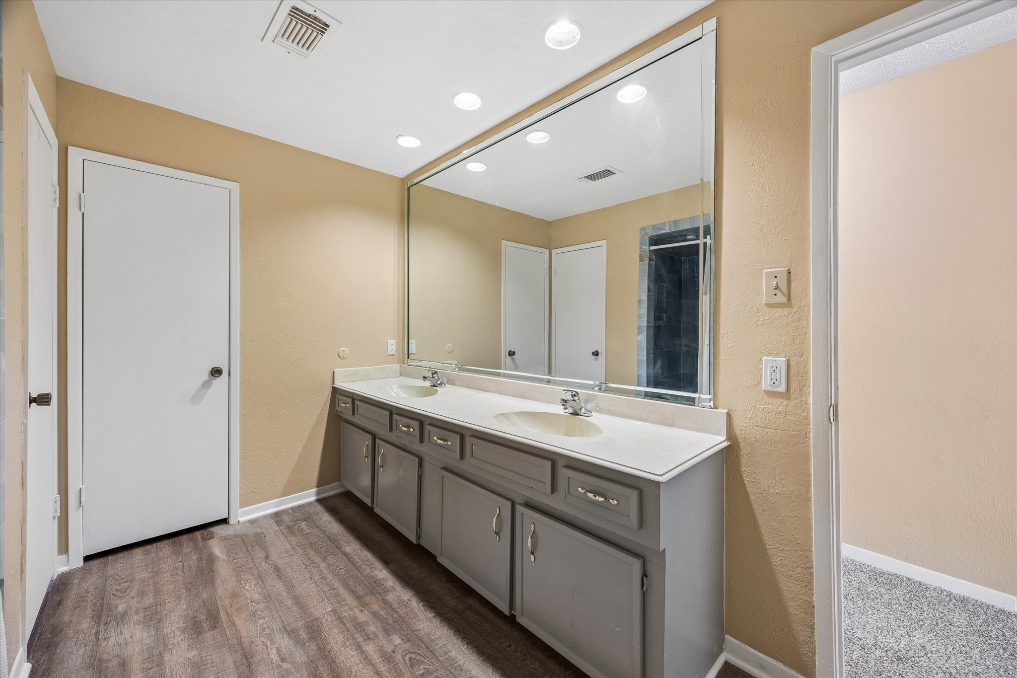 Spacious bathroom featuring a large mirror, sleek vanity with dual sinks, and elegant wood flooring, providing a modern and functional space.