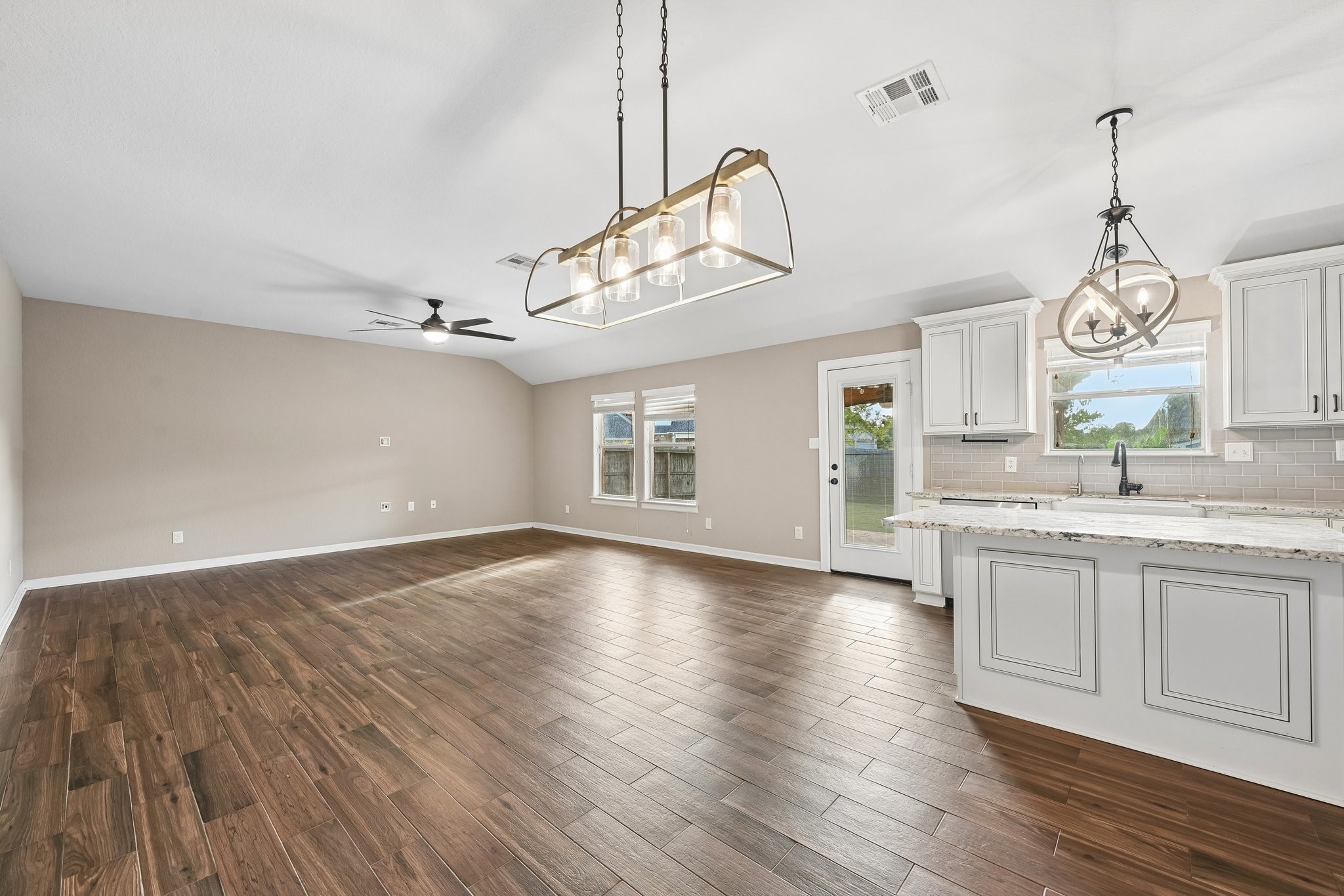 Spacious open-concept living area with elegant wood flooring, modern light fixtures, and a bright kitchen featuring granite countertops and white cabinetry. Natural light floods through large windows and a glass patio door.