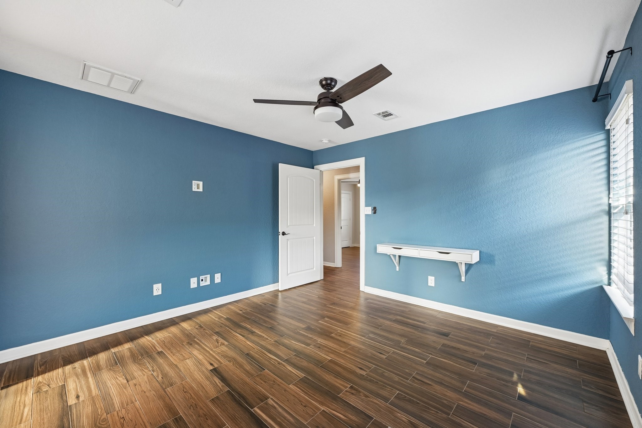 Spacious room with rich wooden flooring and calming blue walls, featuring a modern ceiling fan and ample natural light from large windows.