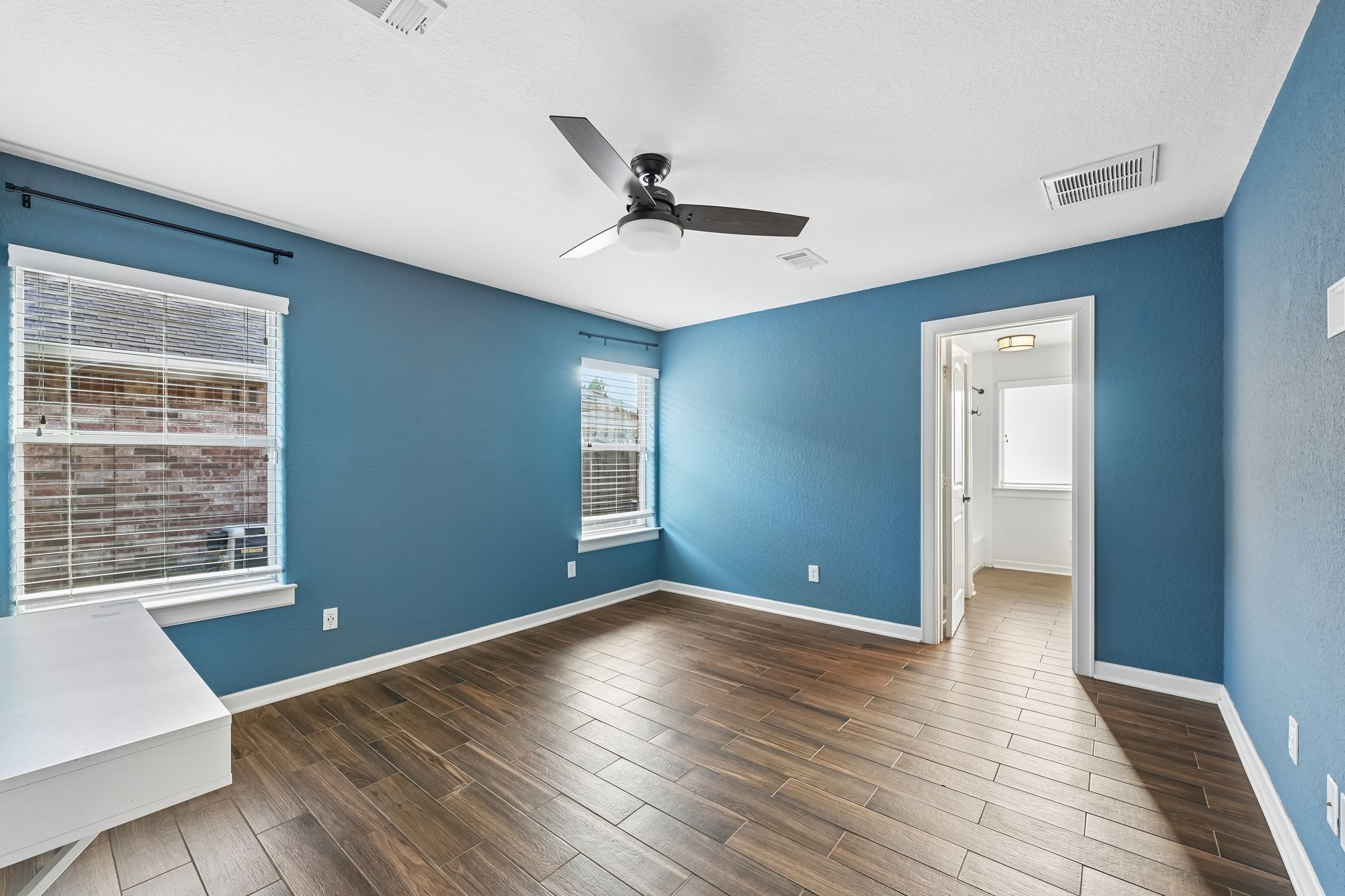 Bright, spacious room featuring elegant wood-look flooring, soothing blue walls, and a modern ceiling fan. Abundant natural light flows through large windows, leading to an adjoining bathroom.