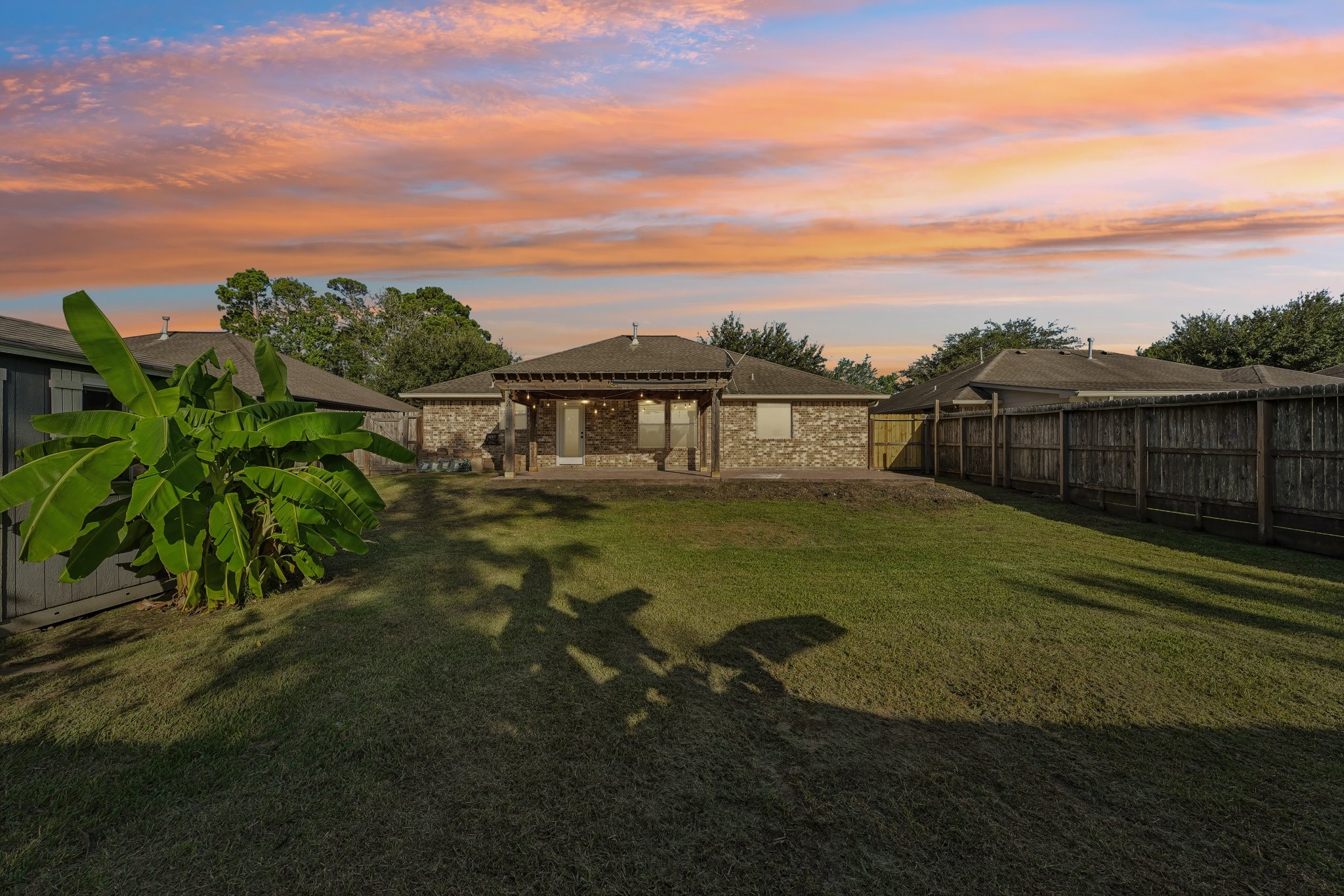 Spacious backyard with well-maintained lawn, privacy fence, and a charming brick exterior under a stunning sunset sky.
