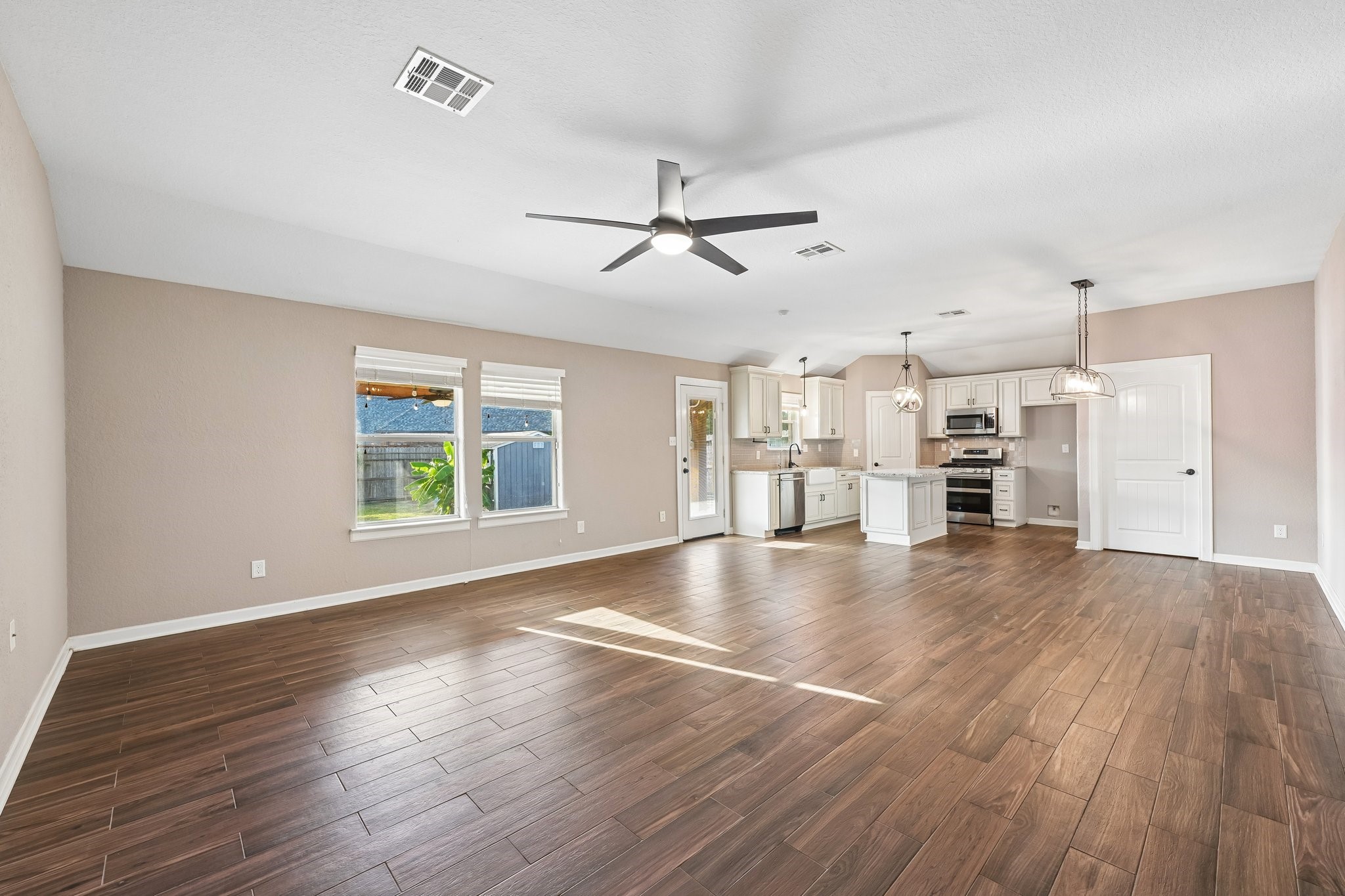 Spacious open-concept living area with elegant wood-look flooring, modern ceiling fan, and a stylish kitchen featuring white cabinetry, stainless steel appliances, and pendant lighting.