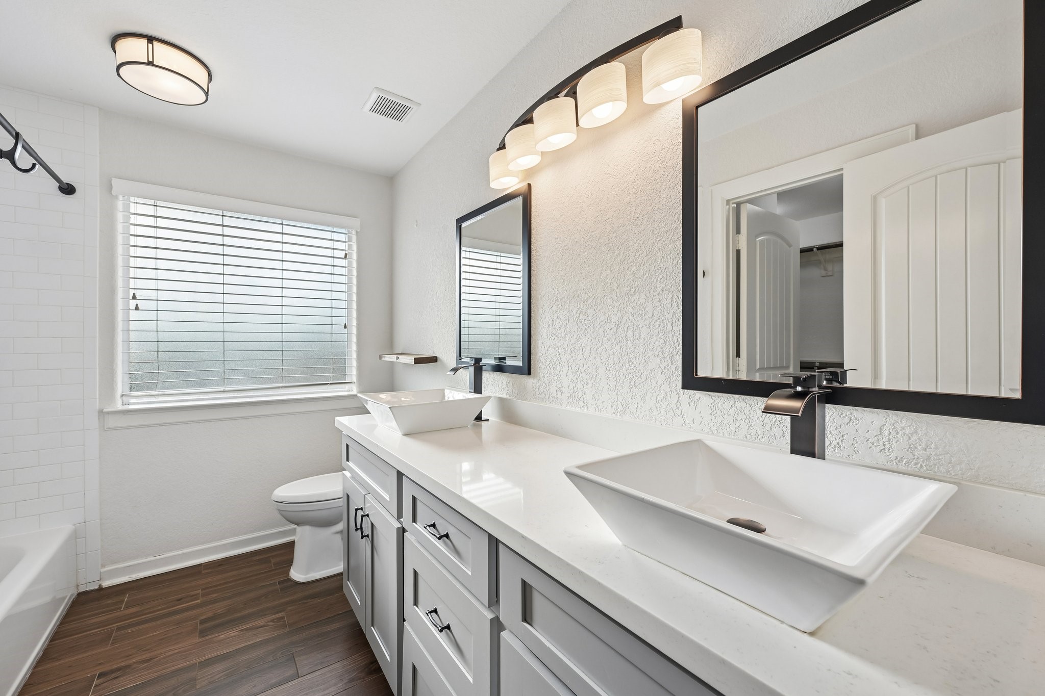 Bright and modern bathroom featuring dual vessel sinks, sleek black fixtures, and elegant lighting, complemented by wood-look tile flooring and a spacious vanity.