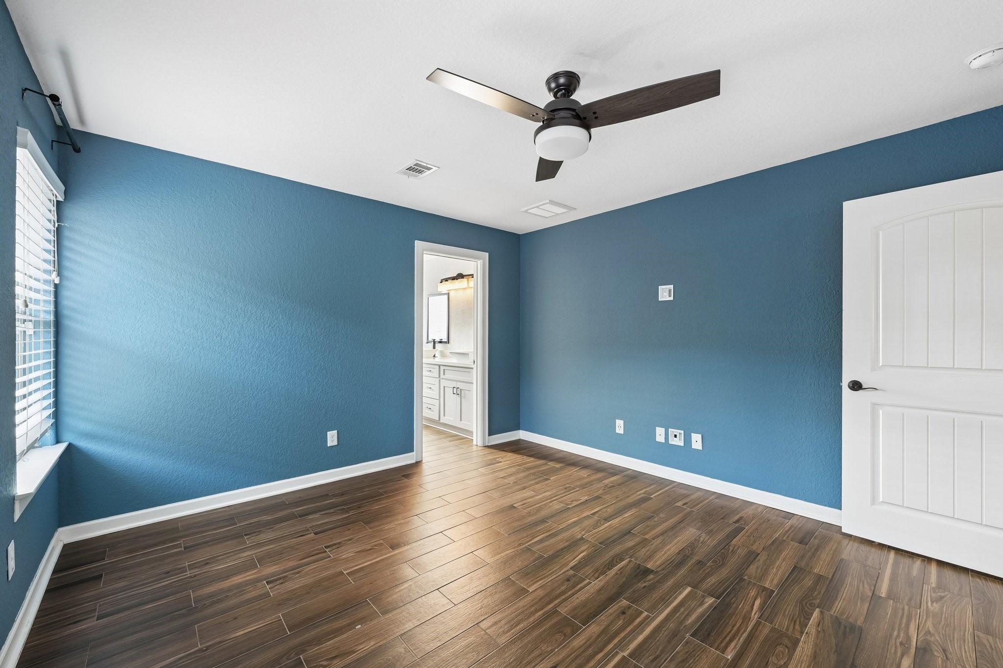 Bright room with rich wood flooring, calming blue walls, and a modern ceiling fan. Natural light streams through large windows, leading to an elegant ensuite bathroom.
