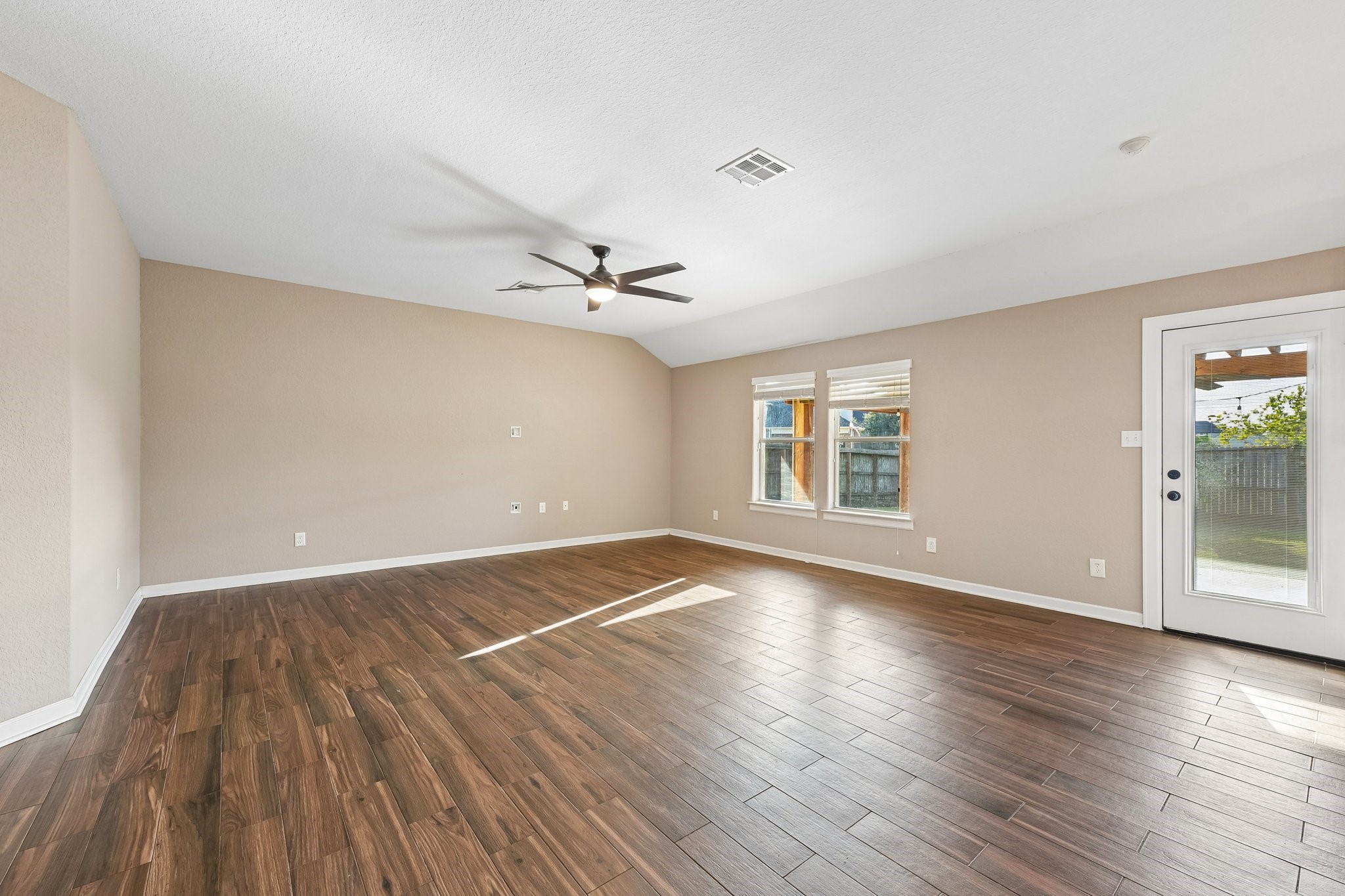 Spacious living area with elegant wood flooring, abundant natural light, and a stylish ceiling fan, leading to a backyard through glass doors.