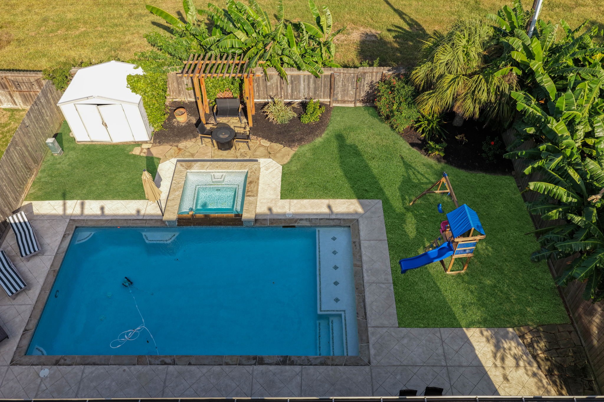 Bird's eye view of the beautifully designed outdoor space featuring a pool and hot tub, pergola seating area, tropical landscaping, and generous green space, all with the added privacy of no back neighbors.