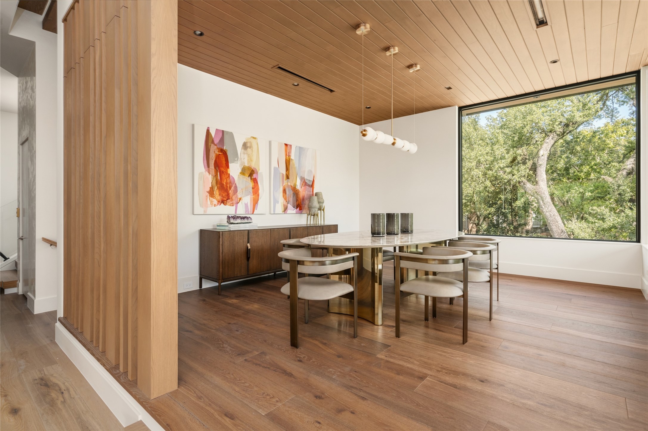 The formal dining room is beautifully defined by a stained tongue-and-groove ceiling, custom wood-slat partitions, and wide-plank European oak flooring, while an oversized picture window frames the treetops like a living work of art.
