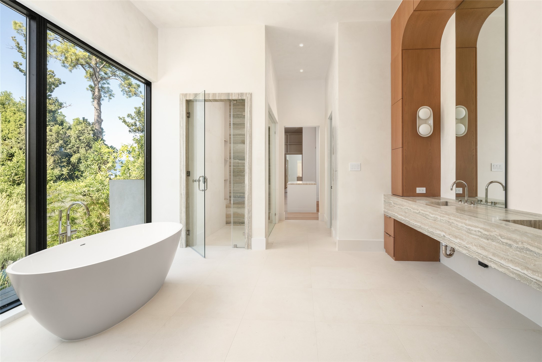 The primary bath pairs a sculptural freestanding soaking tub with a floating double vanity, Silver Travertine finishes, and an oversized picture window overlooking the greenery, creating a serene, spa-inspired setting.