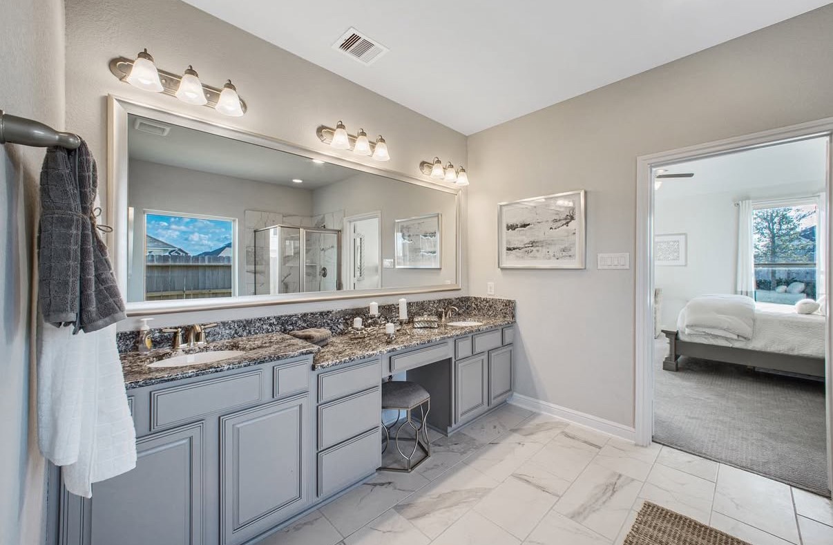 Elegant primary bathroom featuring dual-sinks, granite countertops, a vanity, modern lighting, and a spacious glass-enclosed shower, seamlessly connected to a the cozy primary bedroom.