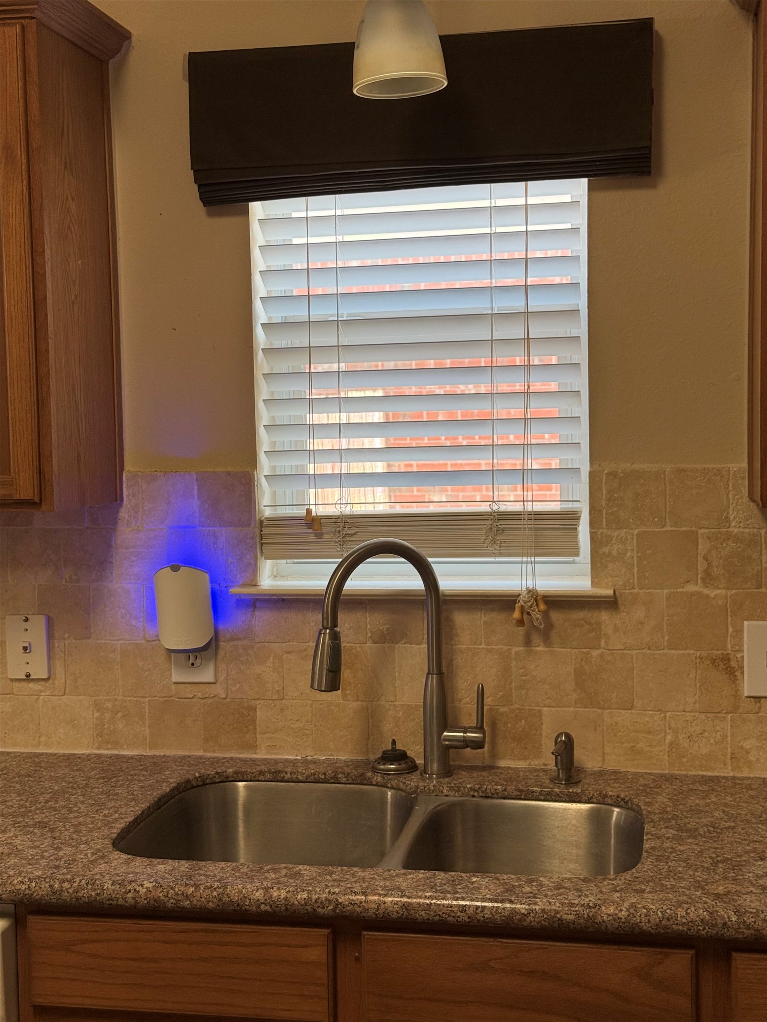 Cozy kitchen corner features a stainless steel double sink under a window with elegant blinds, complemented by warm wooden cabinetry and a stylish backsplash.