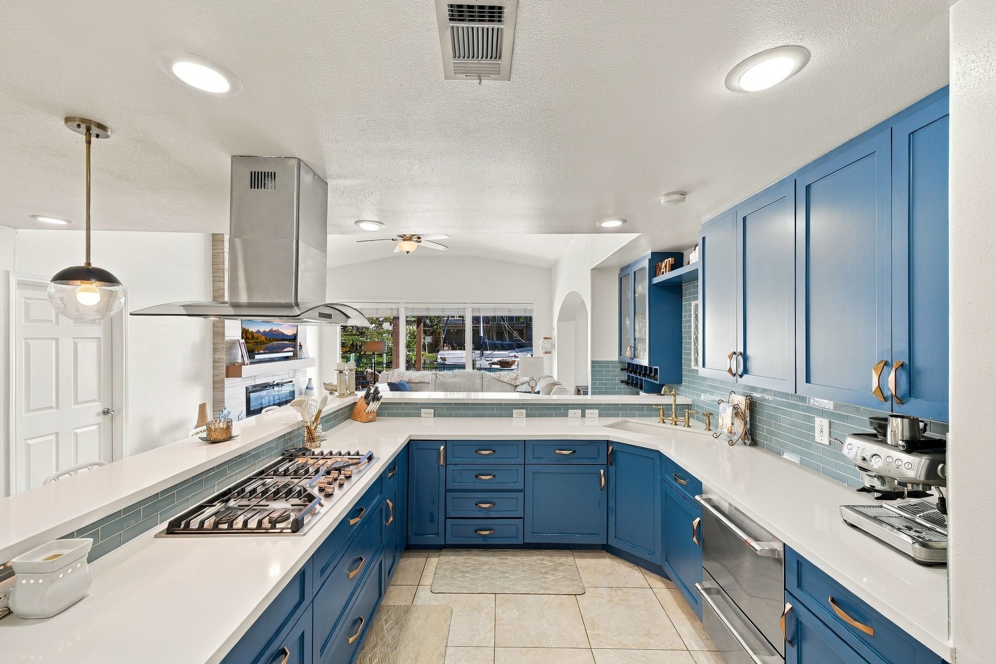 A modern kitchen featuring bold blue cabinetry, sleek countertops, double ovens, and a built-in gas cooktop. The open layout flows into a cozy living area, enhanced by ample natural light.
