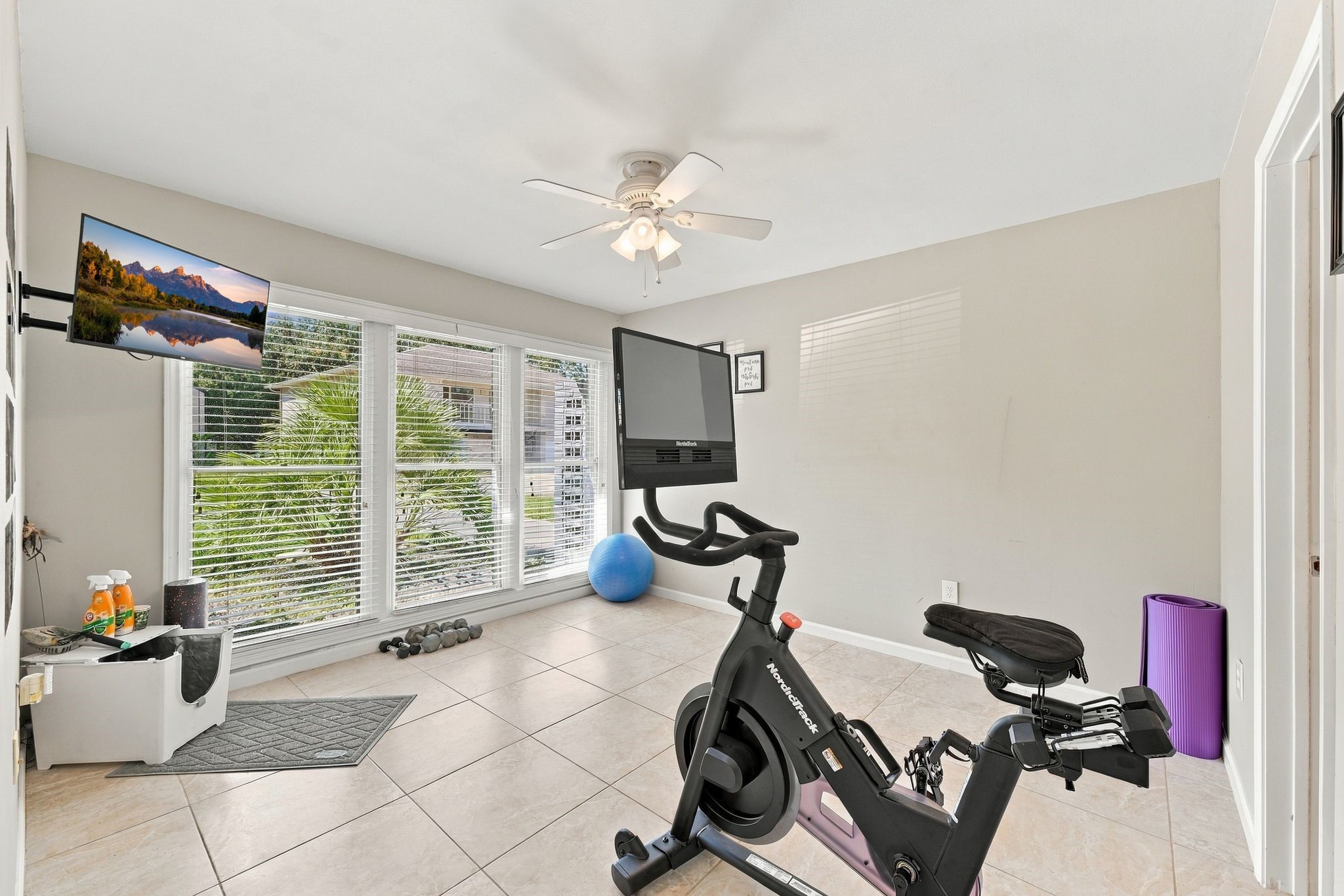 The third bedroom is currently being used as a exercise room with large windows, tiled flooring, and a ceiling fan, offering a perfect space for workouts with a stationary bike and mounted TV.