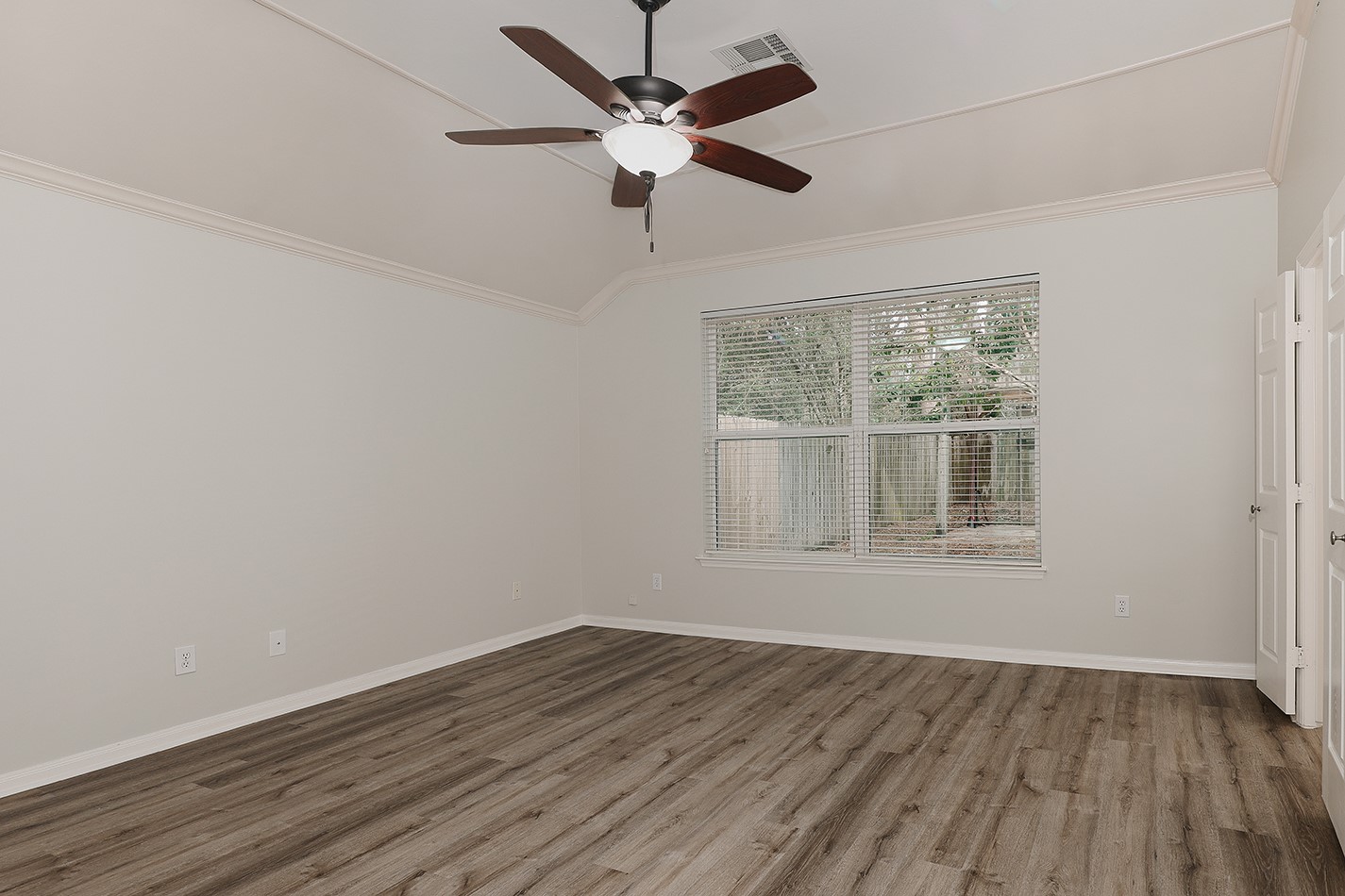 This spacious room features light-colored walls, wood-style flooring, a ceiling fan, and a large window offering natural light and garden views.