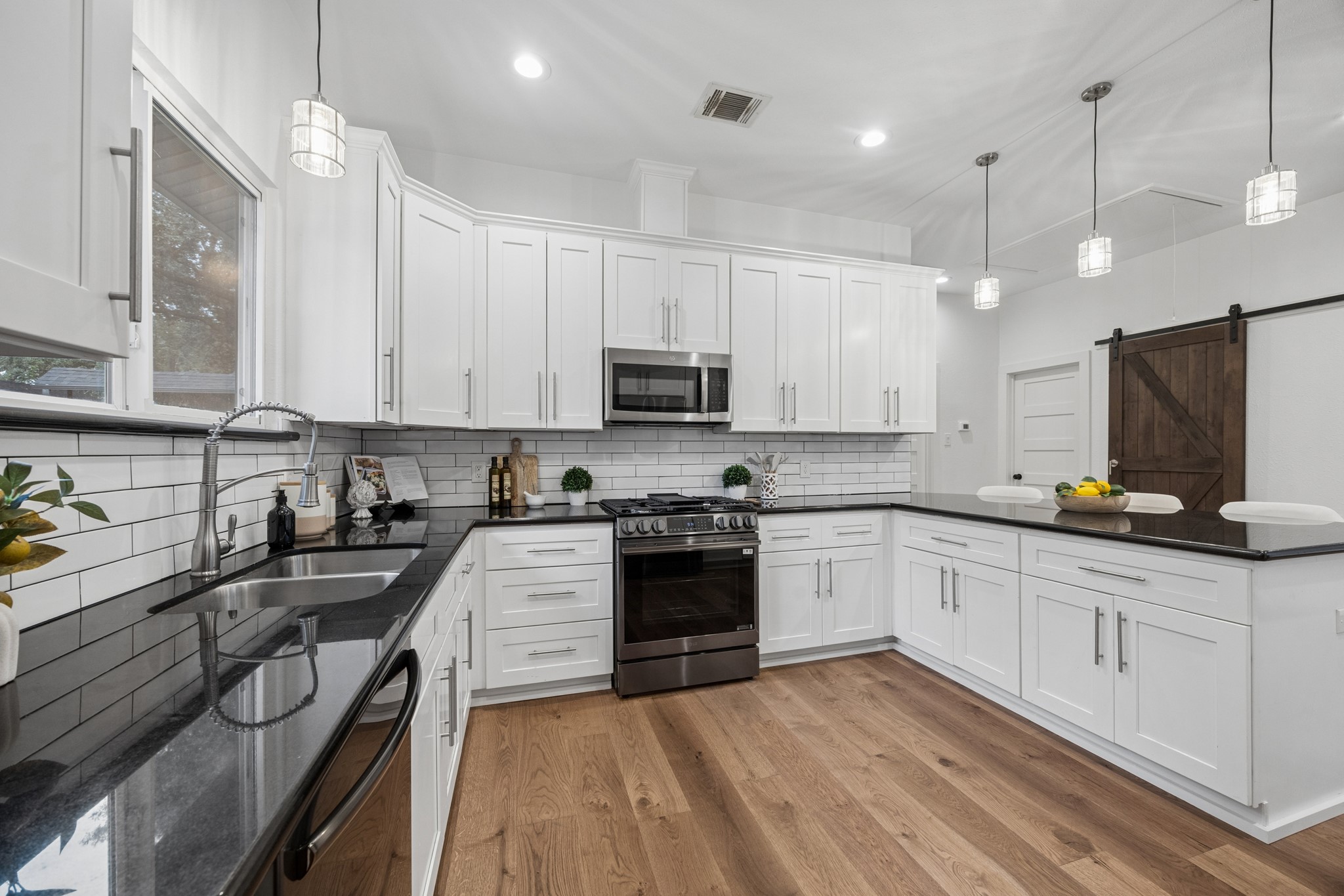 Spacious kitchen with sleek white cabinetry, new stainless steel appliances, and a spacious black countertop. The open layout features a barn door and pendant lighting, complemented by a bright, airy atmosphere and warm wood flooring.
