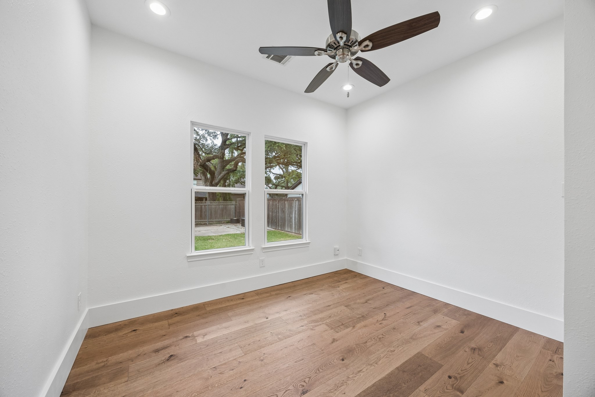 Bedroom featuring elegant hardwood floors, recessed lighting, and a ceiling fan, with large windows offering a view of the lush backyard.