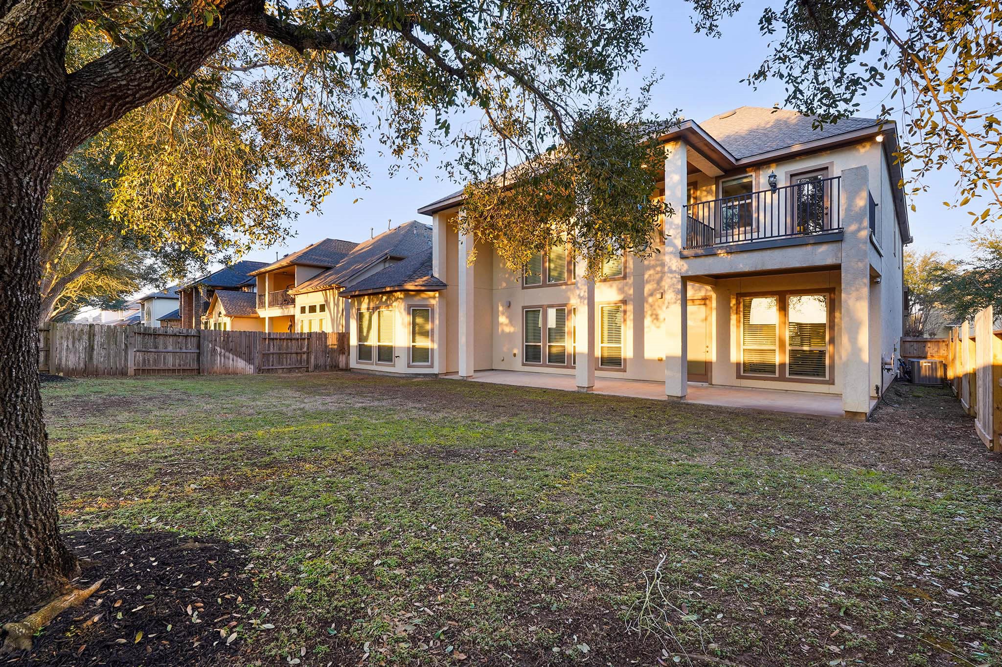 This photo showcases a spacious backyard with a large two-story house. The home features numerous windows for natural light and a balcony on the second floor. The yard is partially shaded by mature trees, offering a comfortable outdoor space.