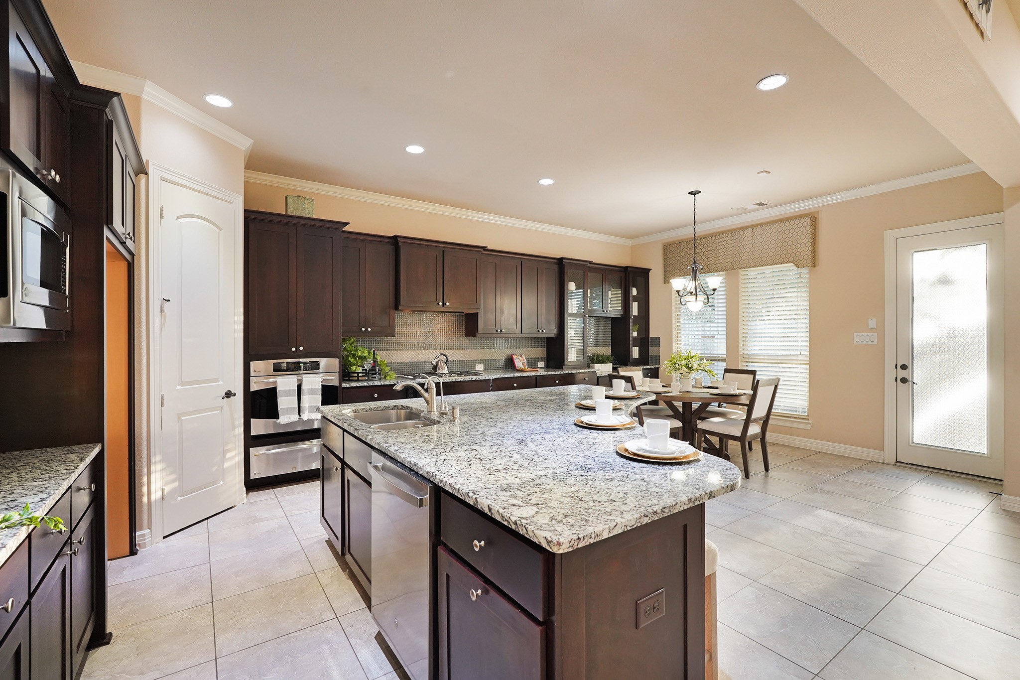 This spacious kitchen features dark wood cabinetry, a large granite island with seating, stainless steel appliances, and a bright dining area with natural light from large windows and a glass door.