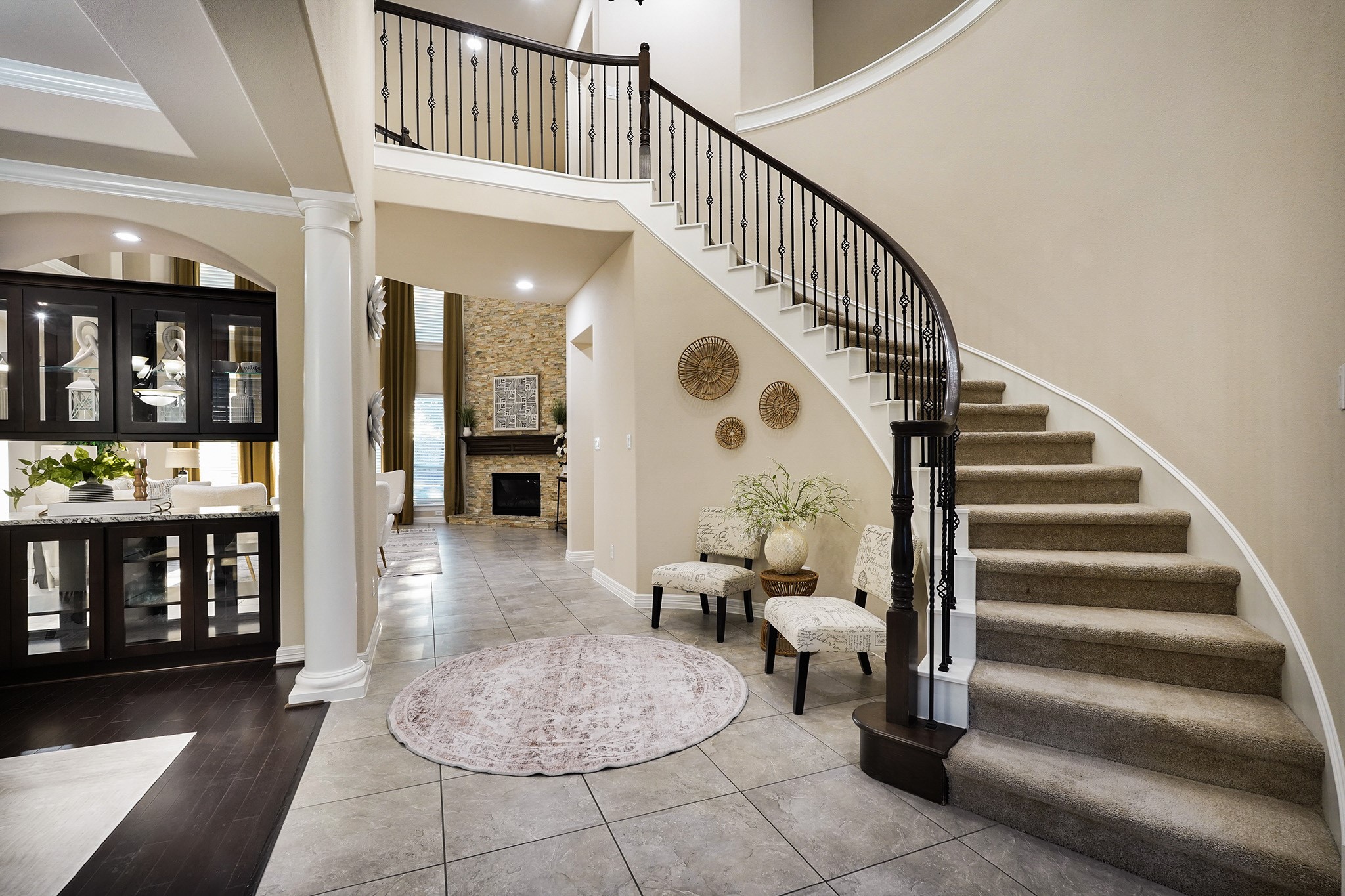 This home features a grand, curved staircase with wrought iron railings, leading to an open, airy foyer. The space is adorned with neutral tones and tiled flooring.