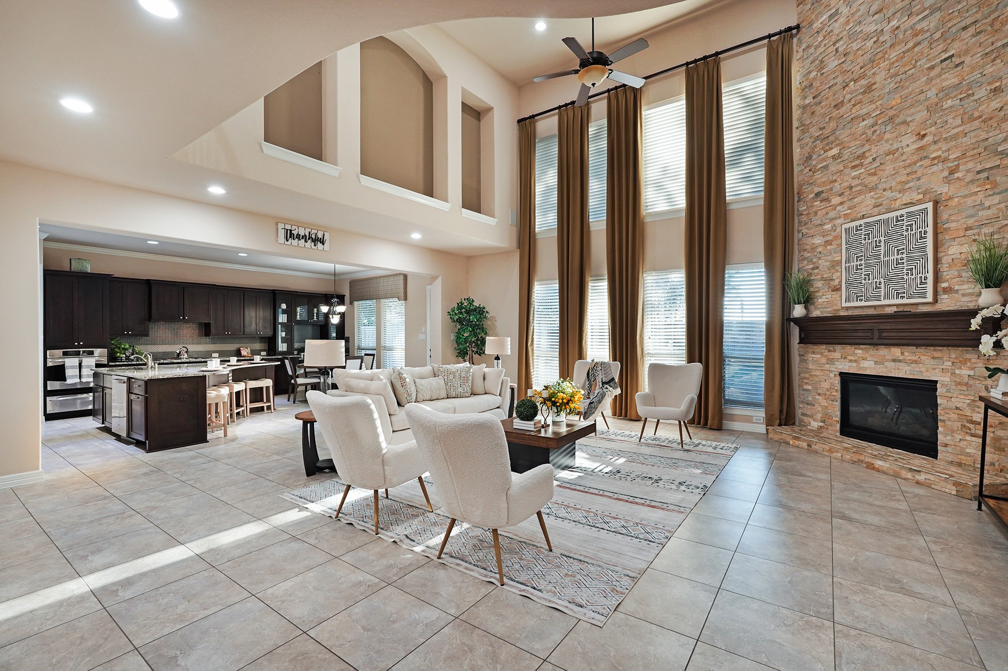 The large family area features high ceilings, large windows with elegant drapes, and a cozy stone fireplace. It flows seamlessly into an open-concept kitchen with dark cabinetry and a central island, perfect for entertaining.