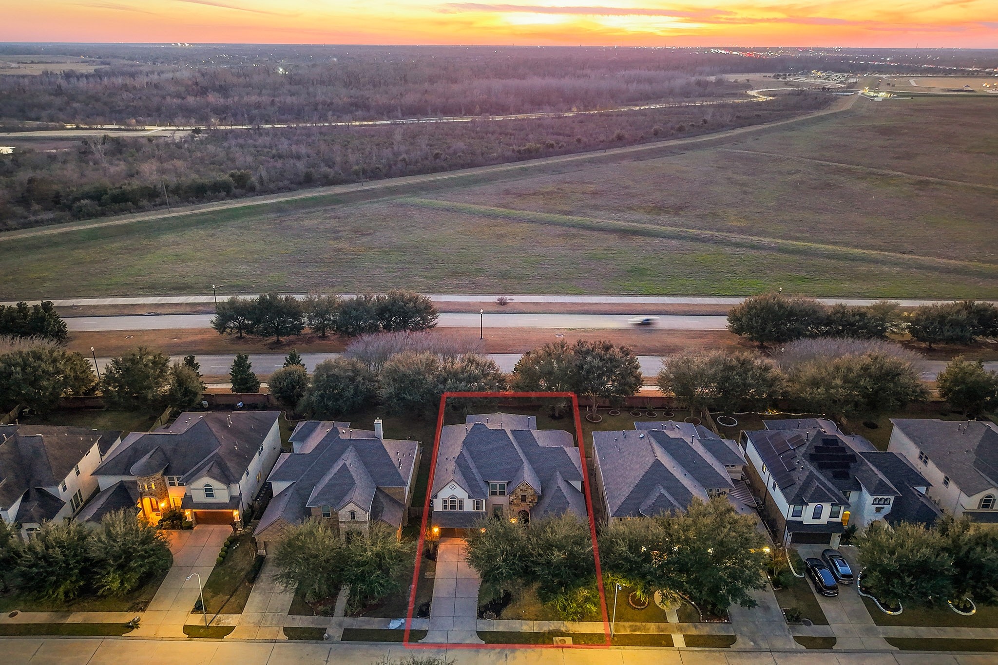 This aerial photo showcases a row of suburban homes with well-maintained lawns and driveways, set against the backdrop of an open field, offering a serene and spacious environment. The sunset in the distance adds a warm, inviting glow to the neighborhood.