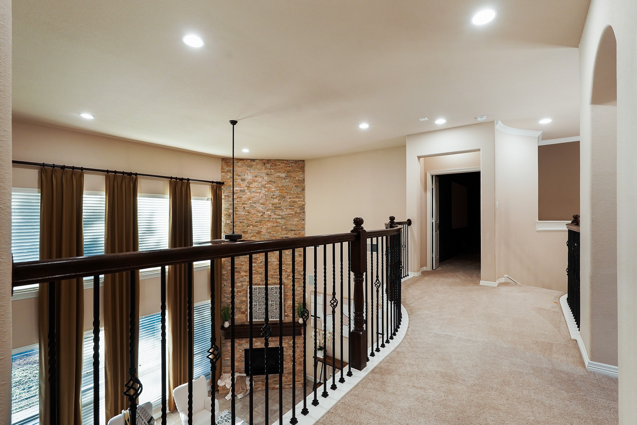 This photo showcases a spacious upstairs hallway featuring elegant railings overlooking a living area with high ceilings and a stone accent wall. The area is well-lit with recessed lighting and has multiple doorways leading to other rooms, offering a warm and inviting ambiance.