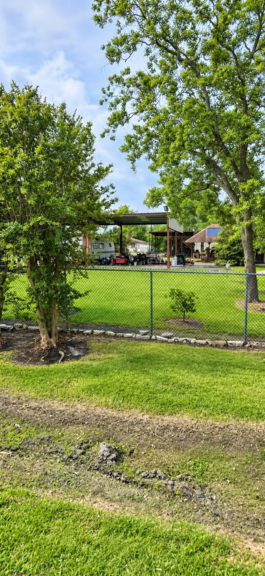 View of the property showcasing the chain-link fencing, mature trees, grassy grounds, and existing covered carport structure