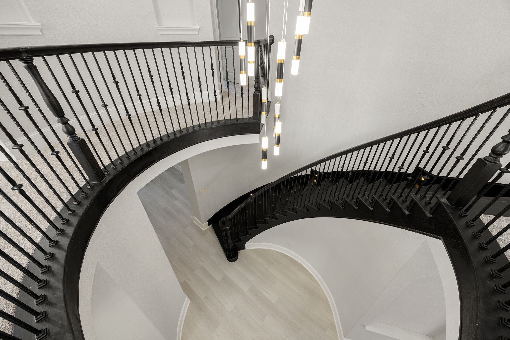 Viewed from above, the sweeping curved staircase becomes a sculptural centerpiece, framed by dark wood handrails, black iron balusters, and a cascading modern light fixture.