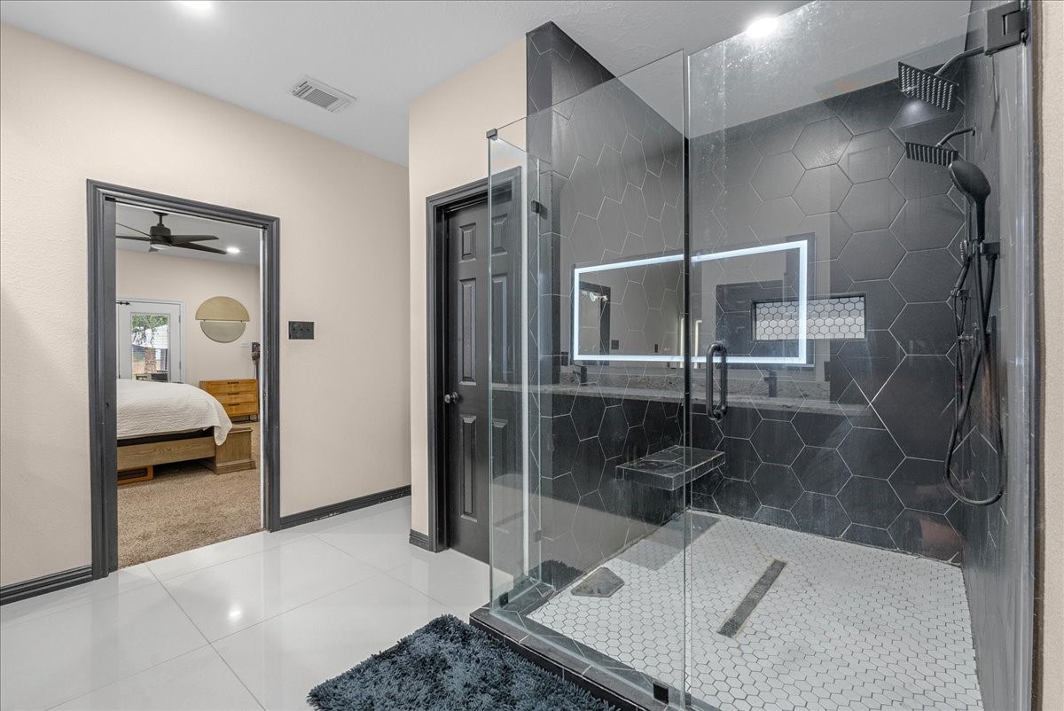 Luxurious primary bathroom featuring a spacious glass-enclosed shower with sleek hexagonal tiles and modern fixtures, complemented by bright, polished flooring. A glimpse into the cozy adjoining bedroom adds to the inviting atmosphere.