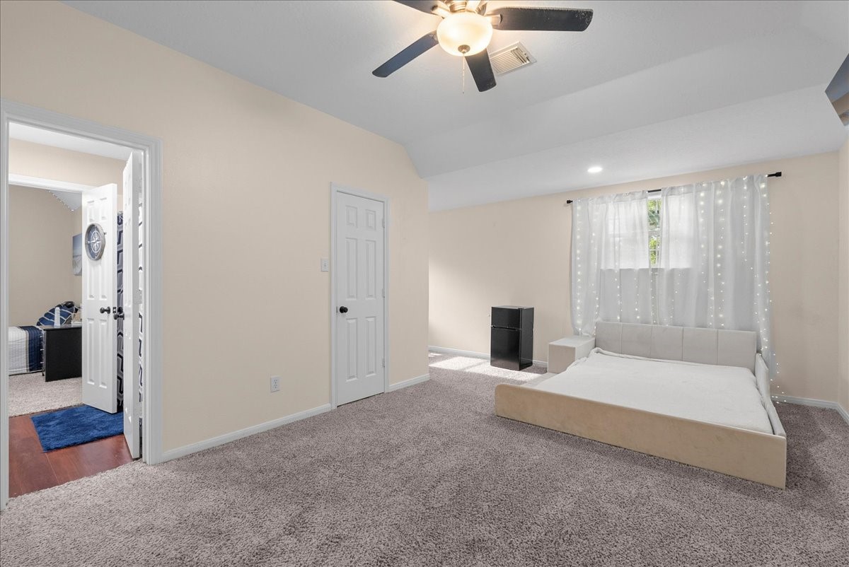 Spacious bedroom with soft carpeting, featuring a ceiling fan and ample natural light from a large window, complemented by a modern bed and neutral decor.