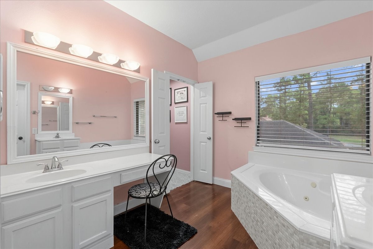 Charming upstairs primary bathroom featuring a spacious vanity with ample lighting, a cozy makeup area, elegant wood like flooring, and a relaxing corner bathtub by a bright window with scenic views.