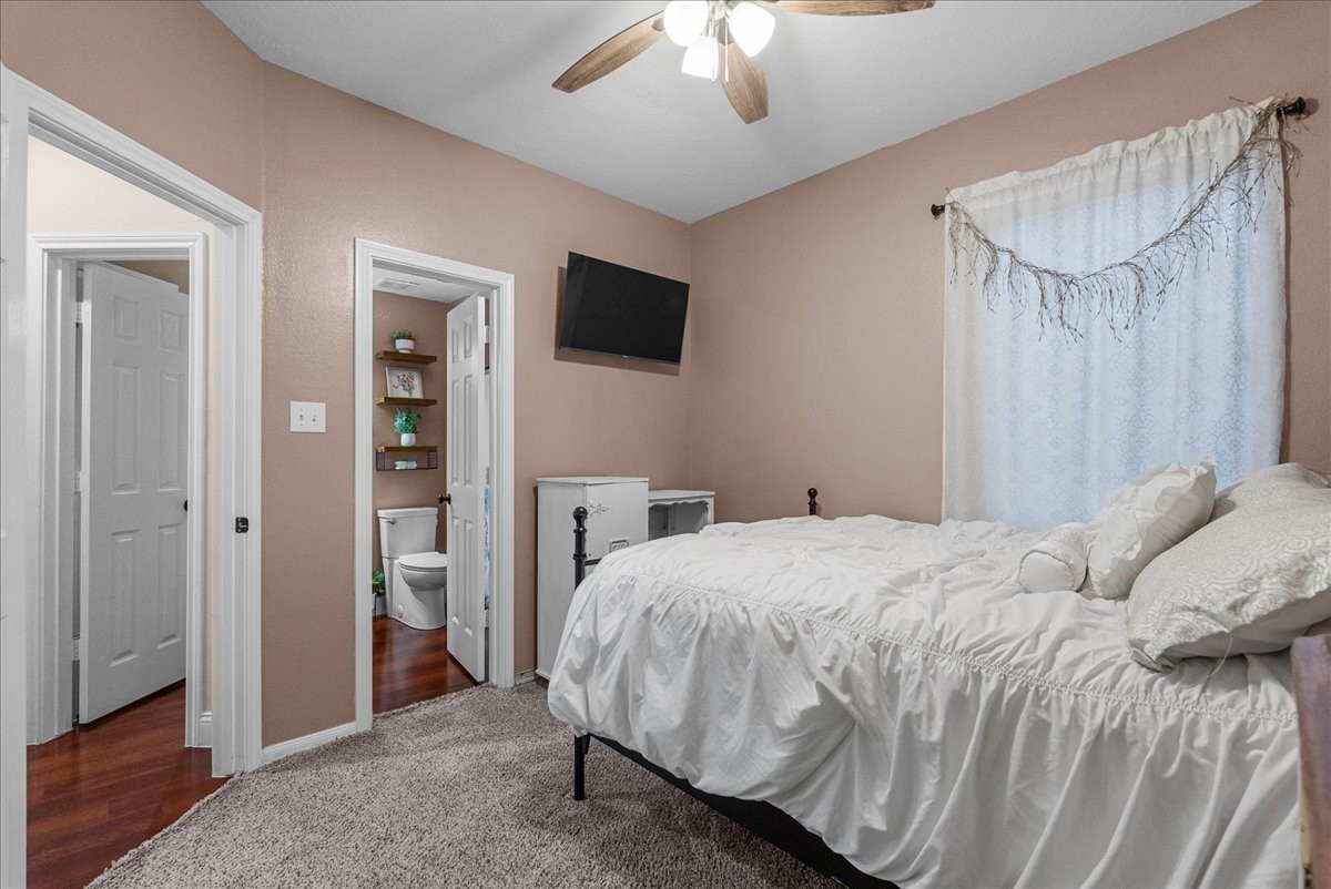 Cozy bedroom with warm tones features a plush carpet, ceiling fan, and wall-mounted TV, seamlessly connected to an ensuite bathroom for added convenience.
