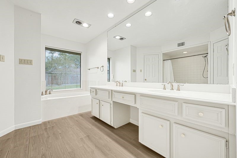 Bathroom features a spacious double vanity with elegant fixtures, a large mirror, and a relaxing soaking tub by the window, all accentuated by stylish wood flooring.