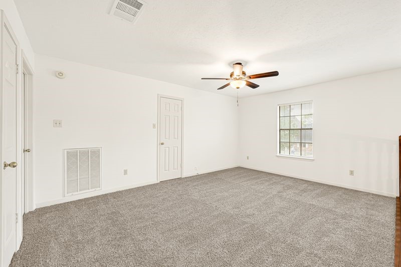 Spacious room with plush carpeting, a ceiling fan, and abundant natural light from a large window, perfect for relaxation.