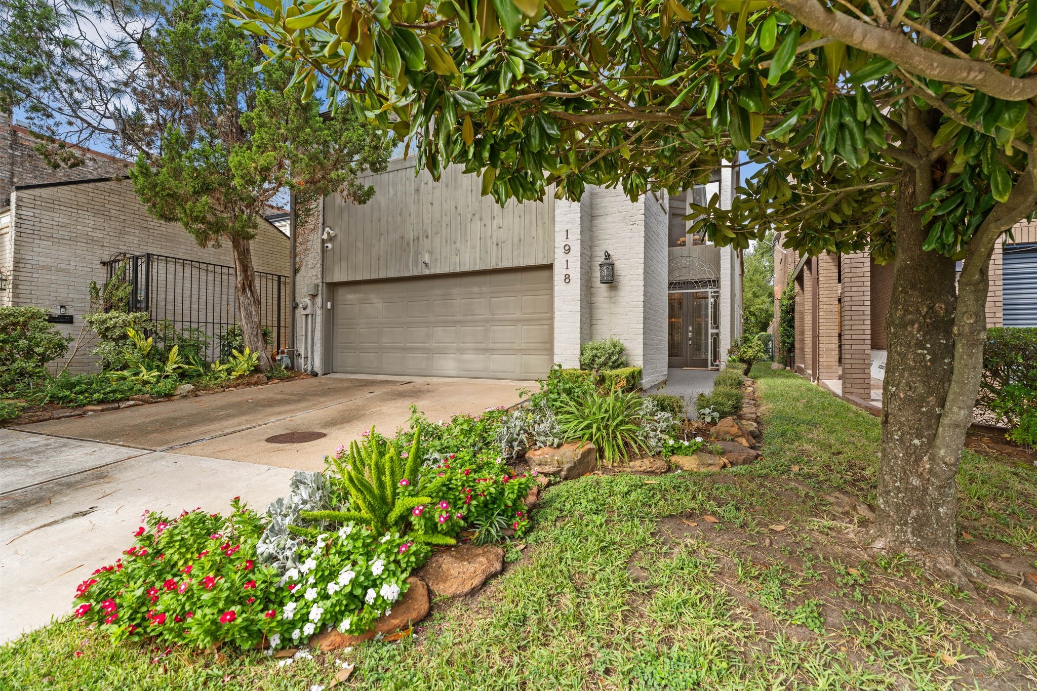 Colorful flower beds and established trees frame the front exterior, with a landscaped walkway leading to the recessed, gated entrance.
