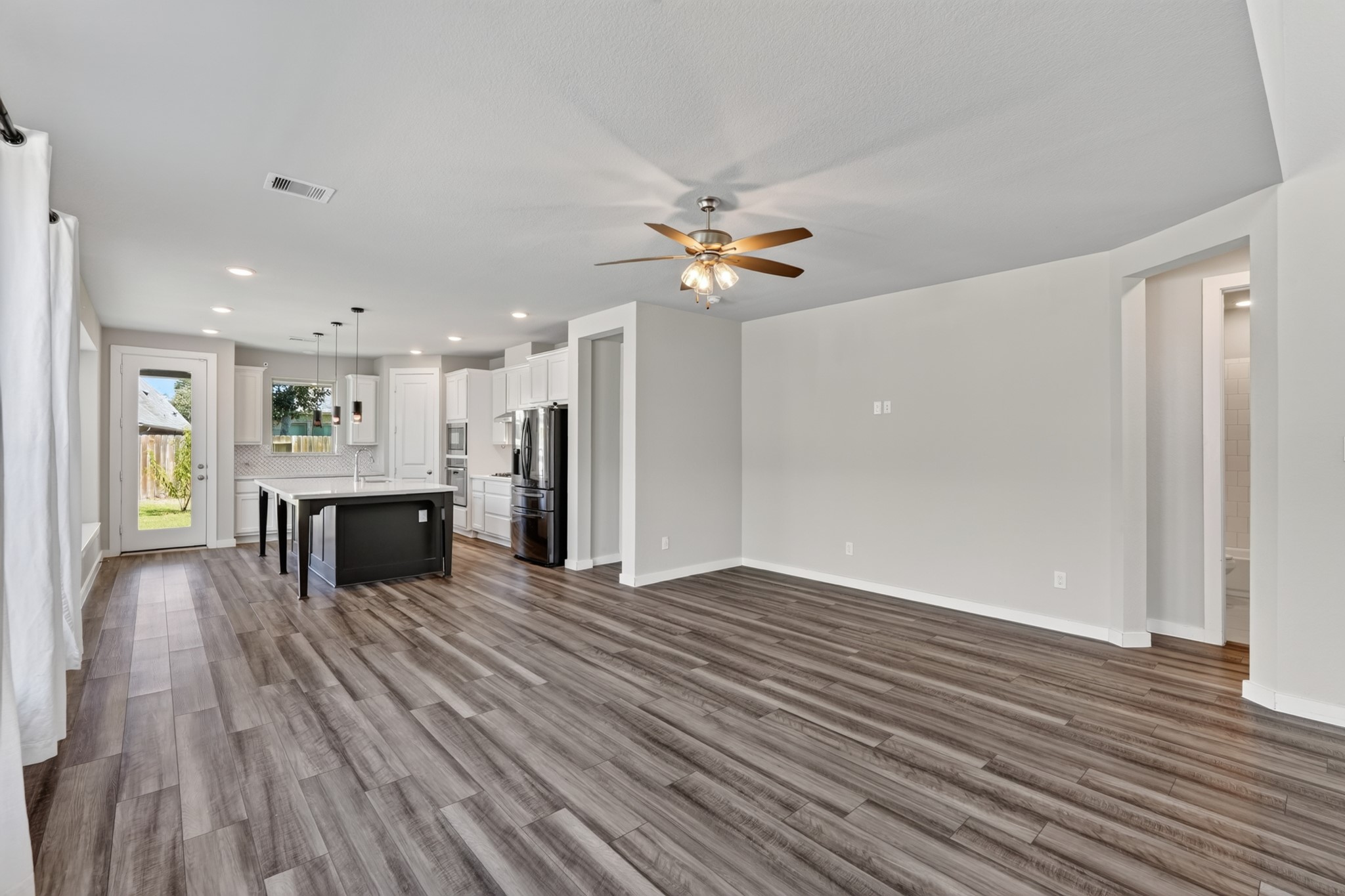 Another angle of the open and airy family room showcases the ceiling fan, dark hardwood flooring, and the seamless open connection to the kitchen and dining areas beyond. This floor plan was designed for real life  and it shows beautifully in every view.