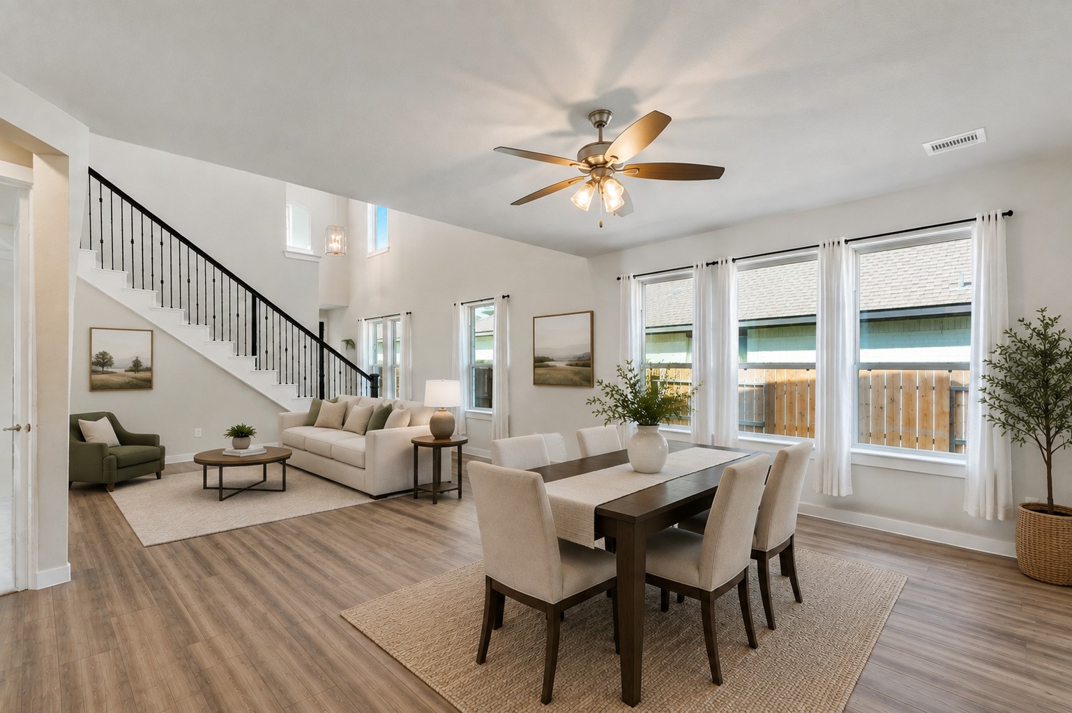 Bright and welcoming family room with a ceiling fan, oversized windows that pour in natural light, and the home's beautiful iron stair railing as a backdrop. Dark hardwood-style flooring flows seamlessly throughout, tying all the downstairs spaces together.