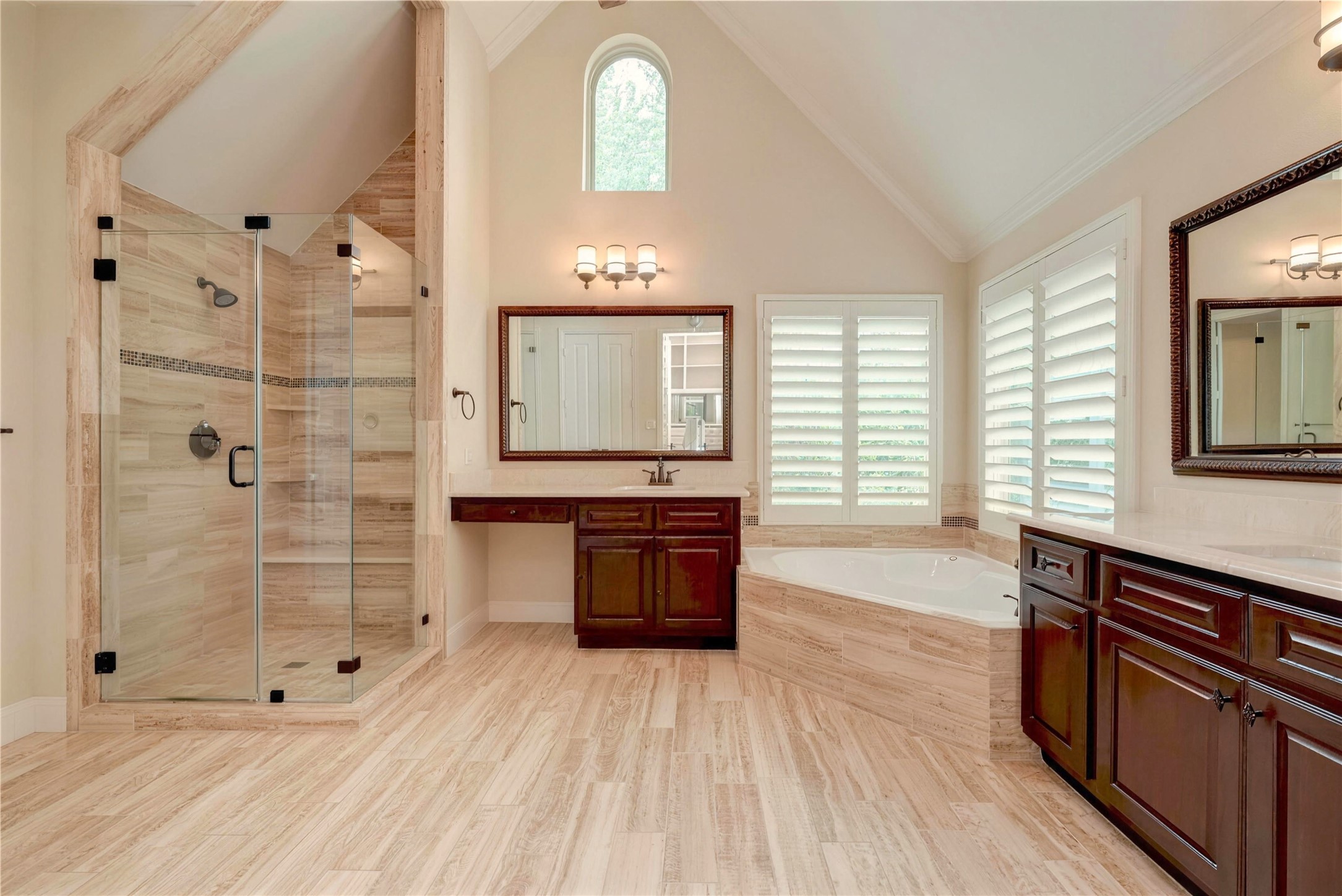 Luxurious bathroom featuring a spacious glass-enclosed shower, elegant soaking tub, and rich wood cabinetry, all illuminated by natural light from a high, arched window.