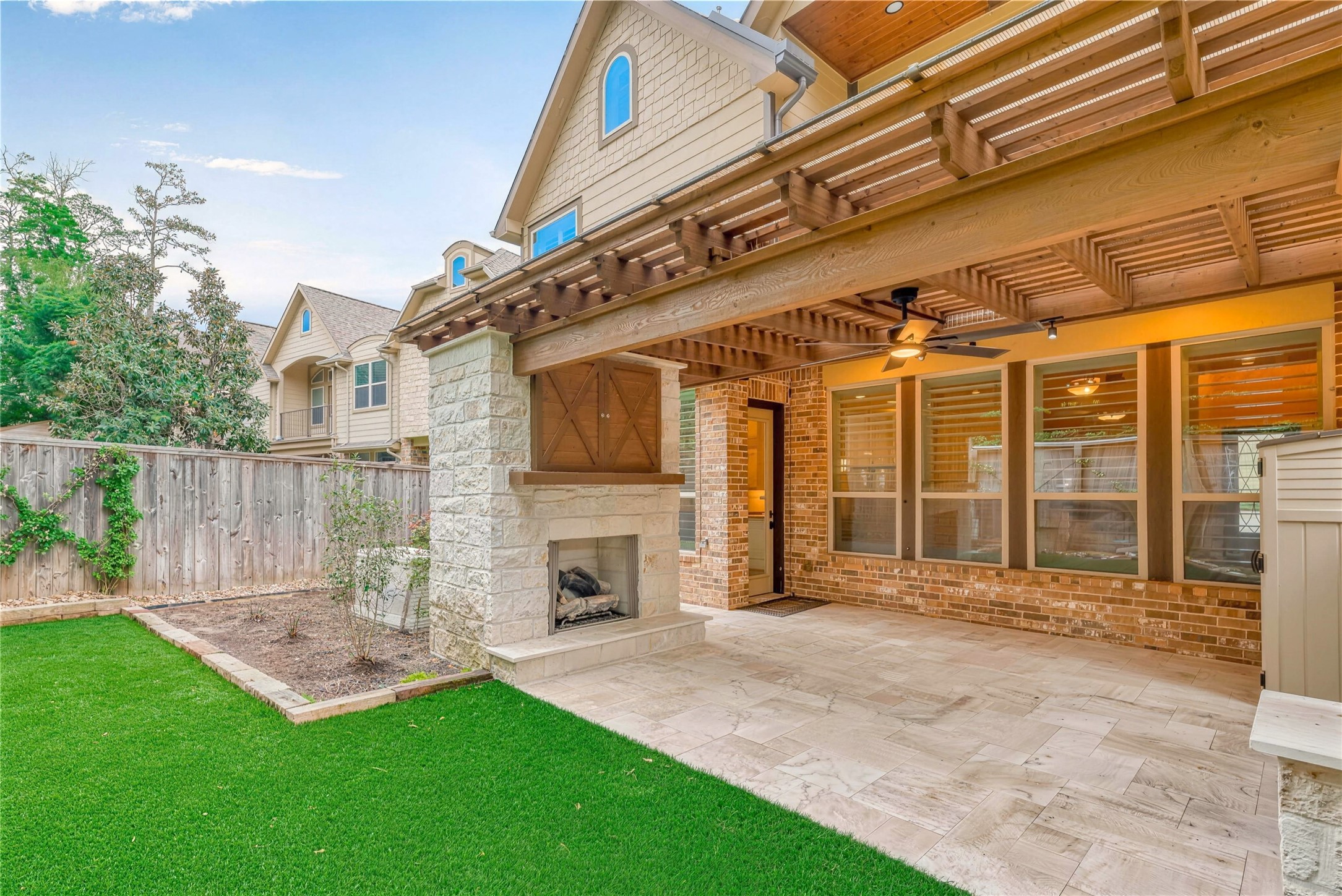 Charming outdoor living space featuring a cozy stone fireplace under a pergola, complemented by lush artificial turf and elegant tile flooring for seamless indoor-outdoor entertaining.