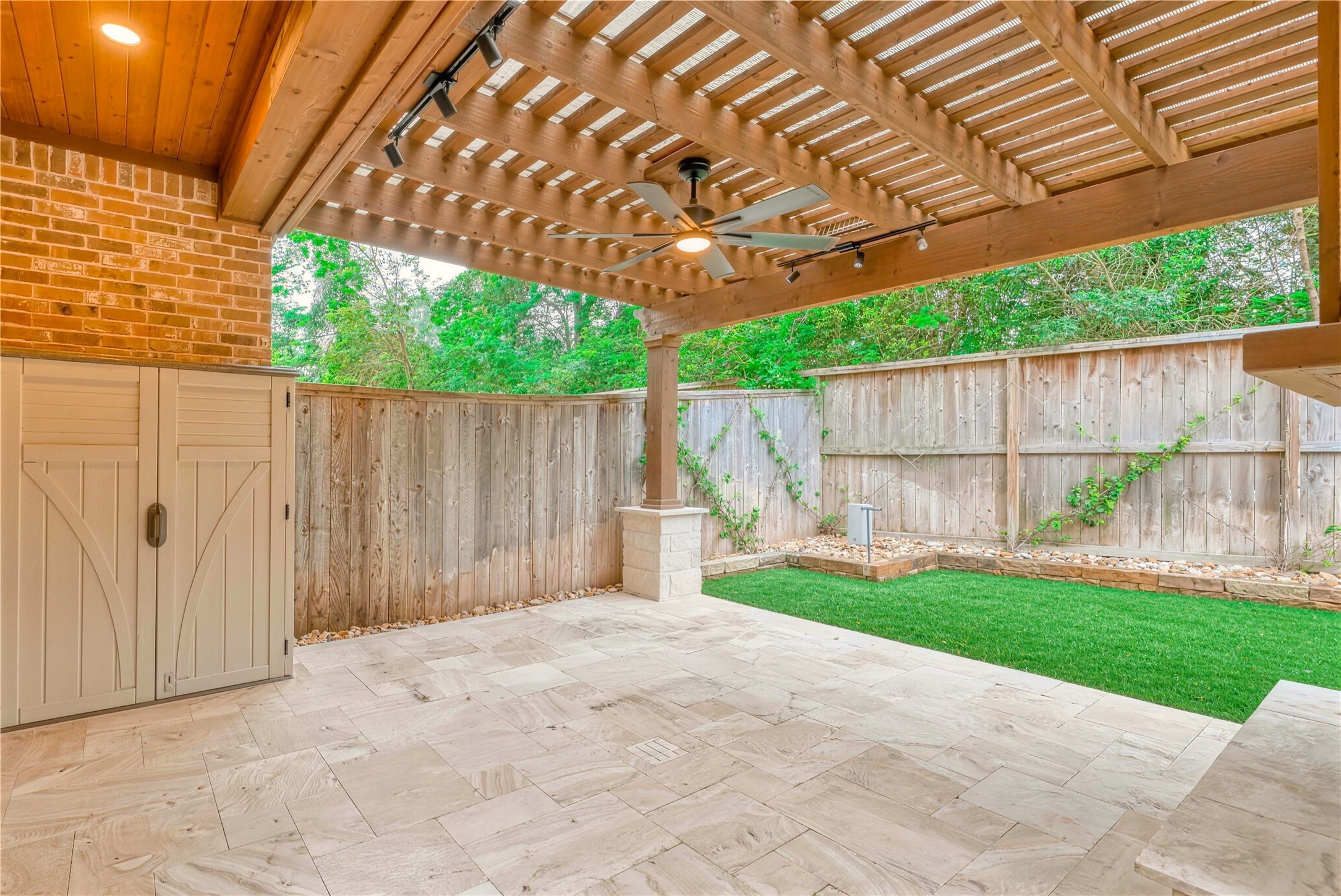 Charming patio with a wooden pergola, ceiling fan, and elegant stone flooring, ideal for outdoor relaxation. The fenced yard offers privacy and a touch of greenery.