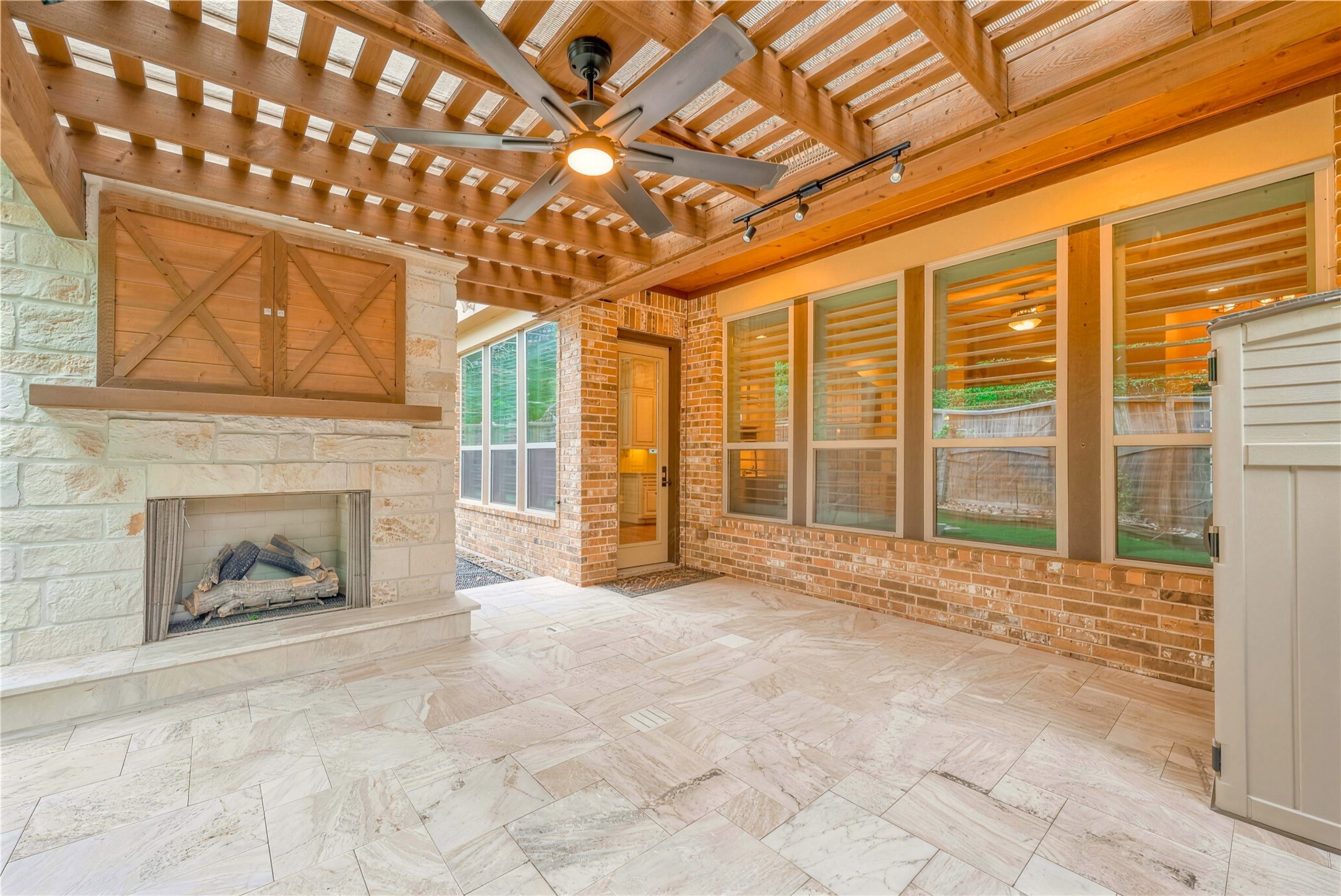 Charming outdoor living space featuring a cozy stone fireplace, sleek ceiling fan, and elegant pergola, perfect for relaxing or entertaining guests.